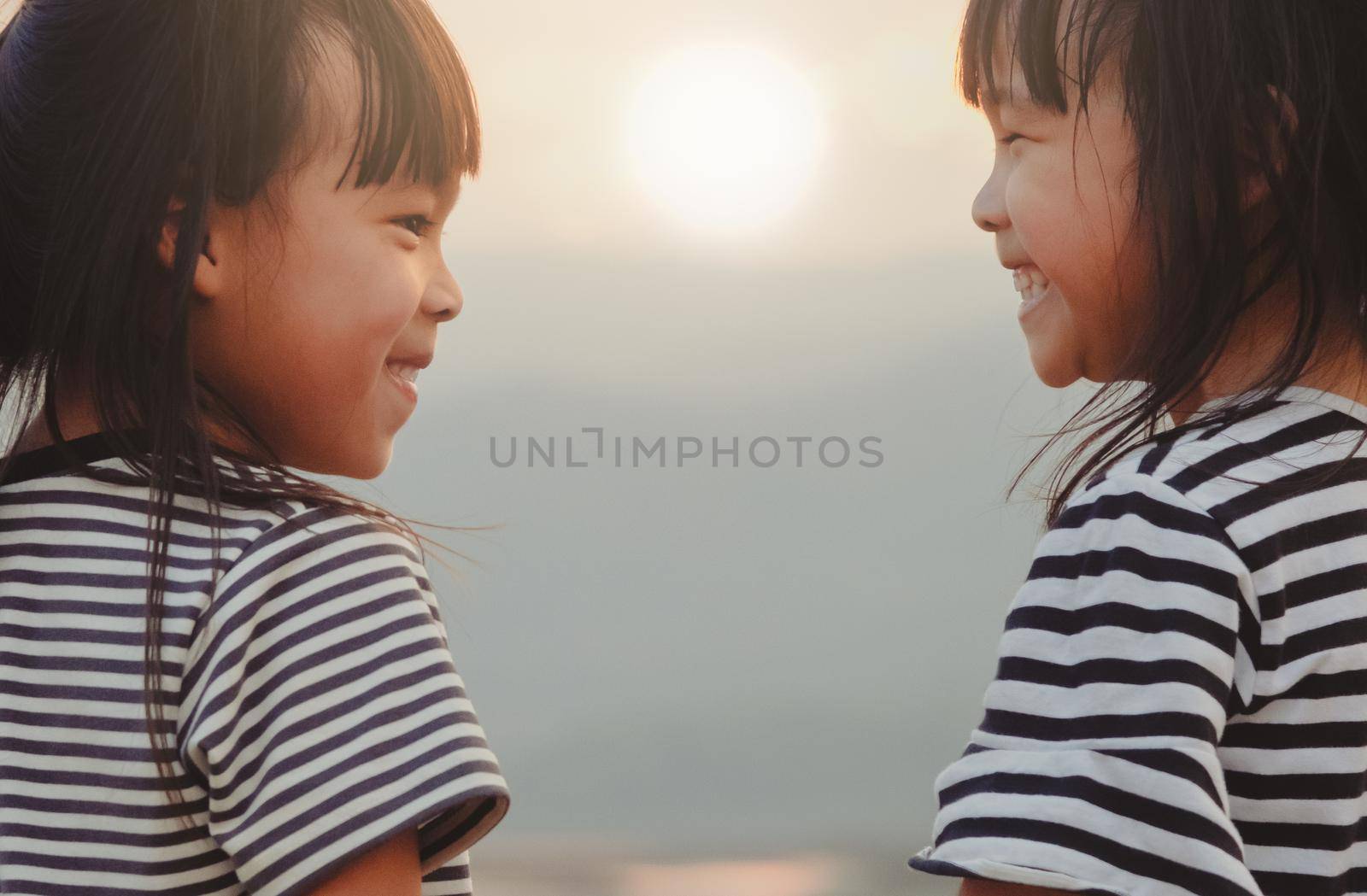 Cute little sisters looking at each other and smiling against the sunset background. The concept of family and sibling love by TEERASAK
