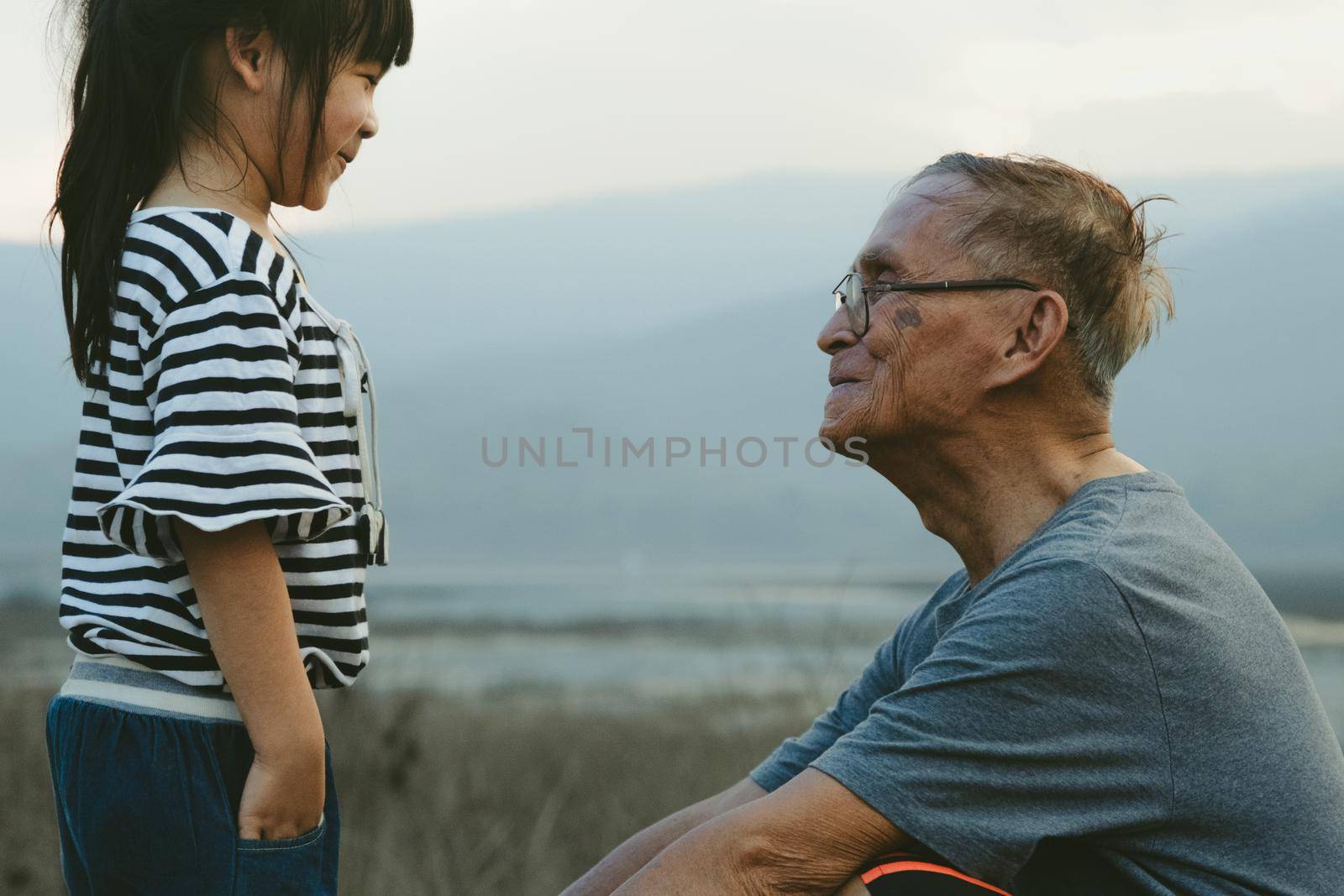 Loving grandfather and little granddaughter look at each other and smile against the sunset background. Family love and spending time outdoors together.