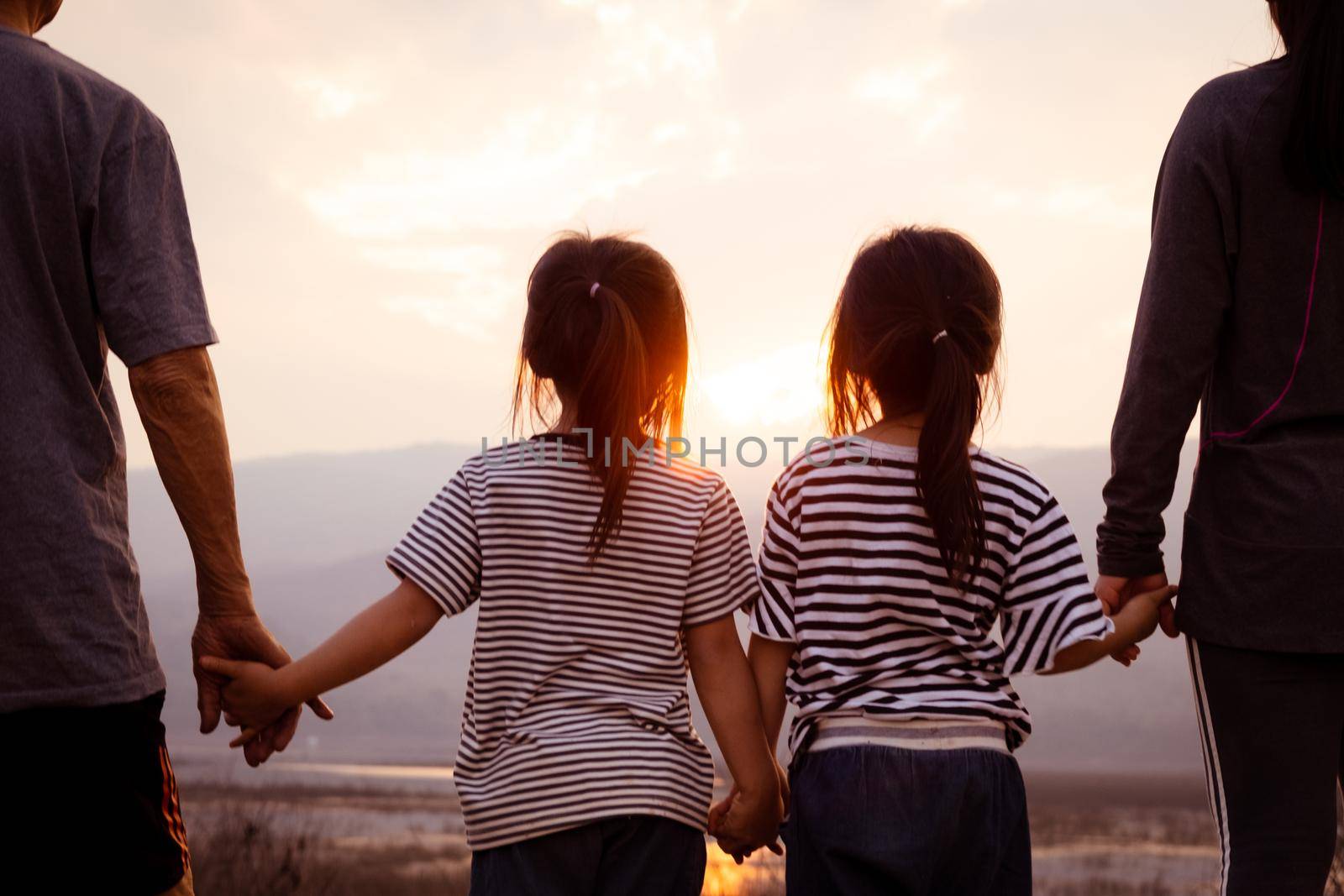 Rear view of family holding hands while standing on the hill and looking at sunset. Happy family enjoying a mountain trip at sunset. by TEERASAK