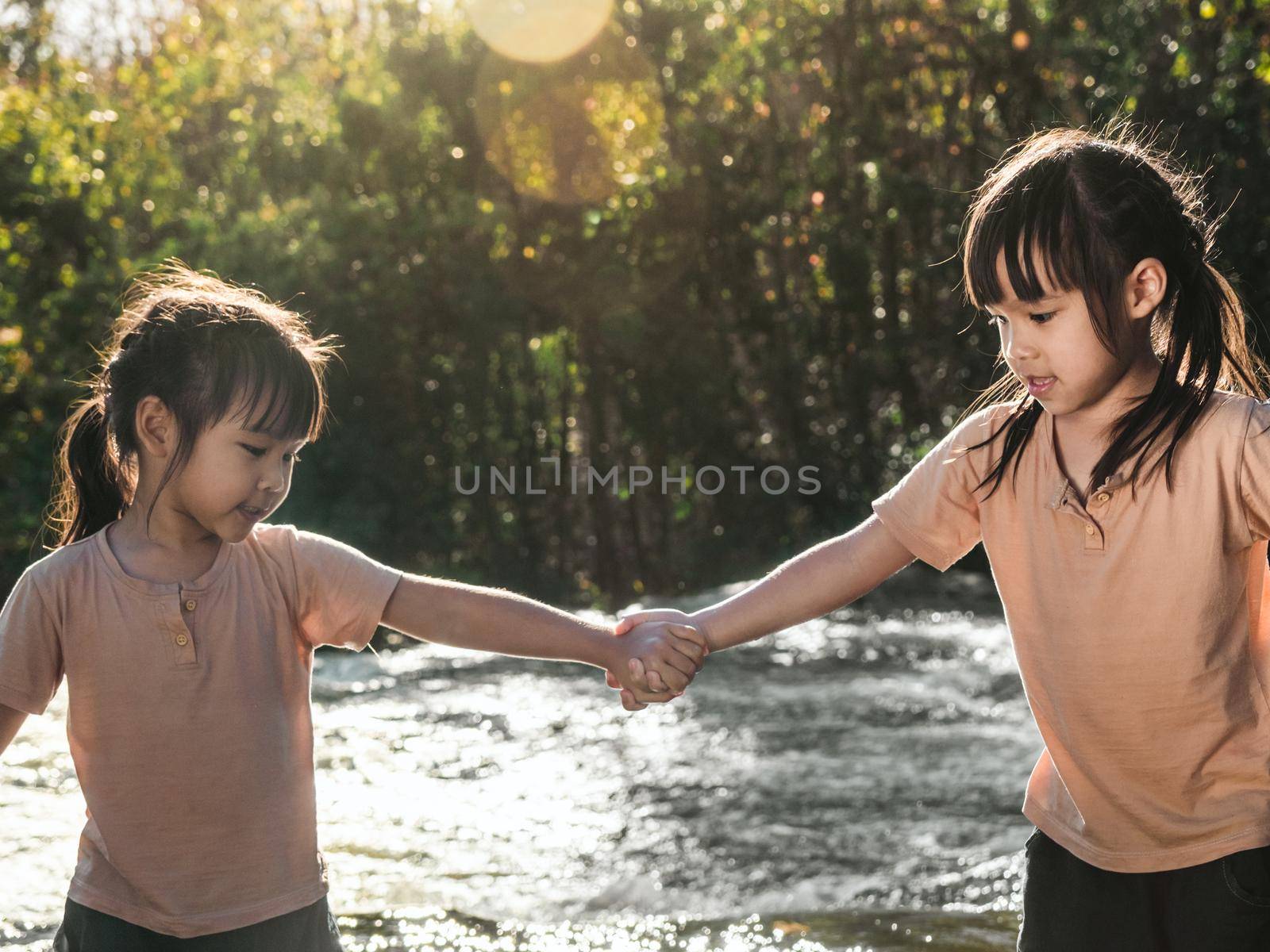Two lovely girls holding hands happily playing in the stream. Asian sibling girls are playing in the waterfall in the forest. Active recreation with children on river in summer. by TEERASAK