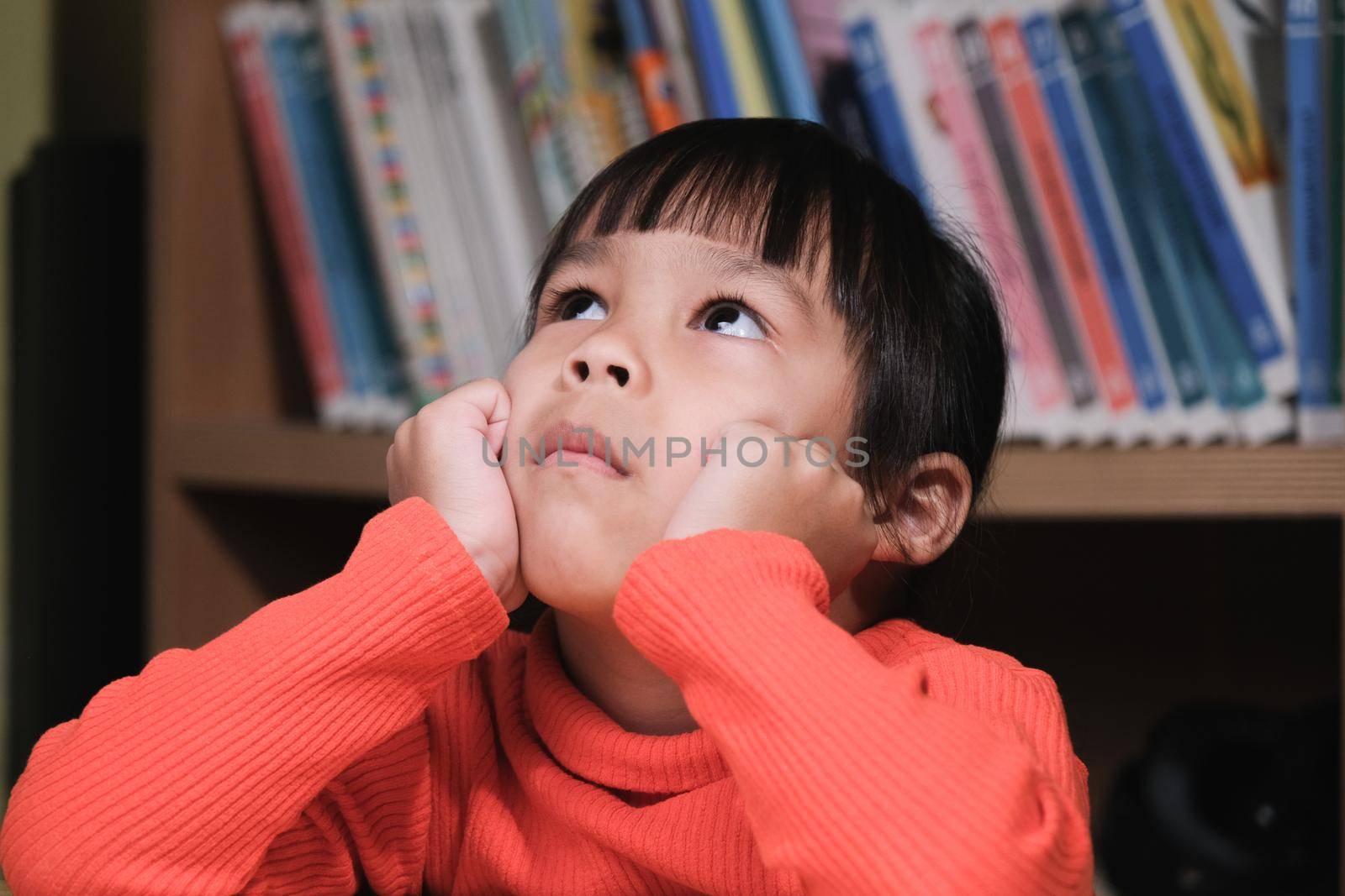 Portrait of cute child looking up and thinking with hands on chin in living room. school child thinking by TEERASAK