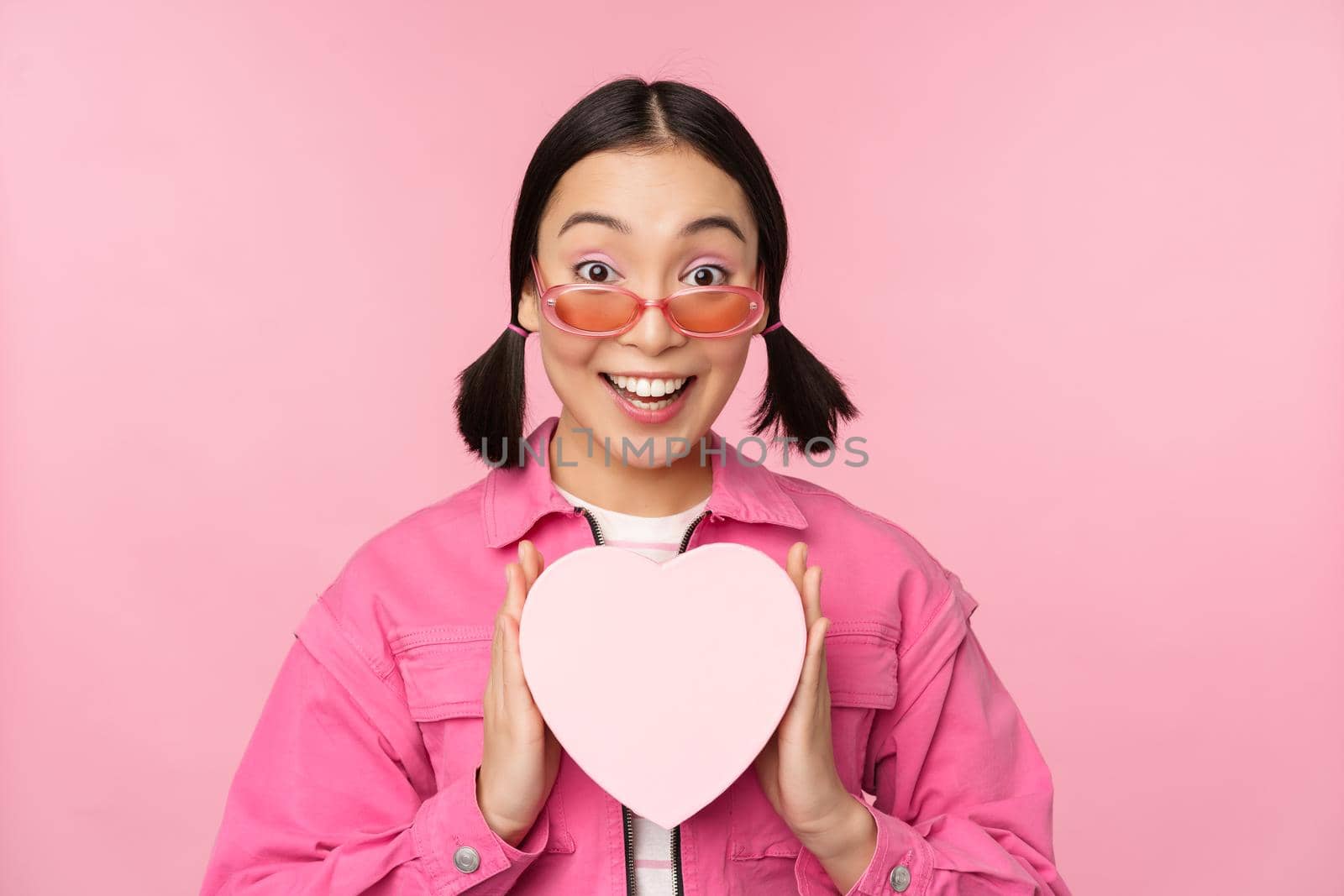 Beautiful asian girl smiling happy, showing heart gift box and looking excited at camera, standing over pink romantic background by Benzoix