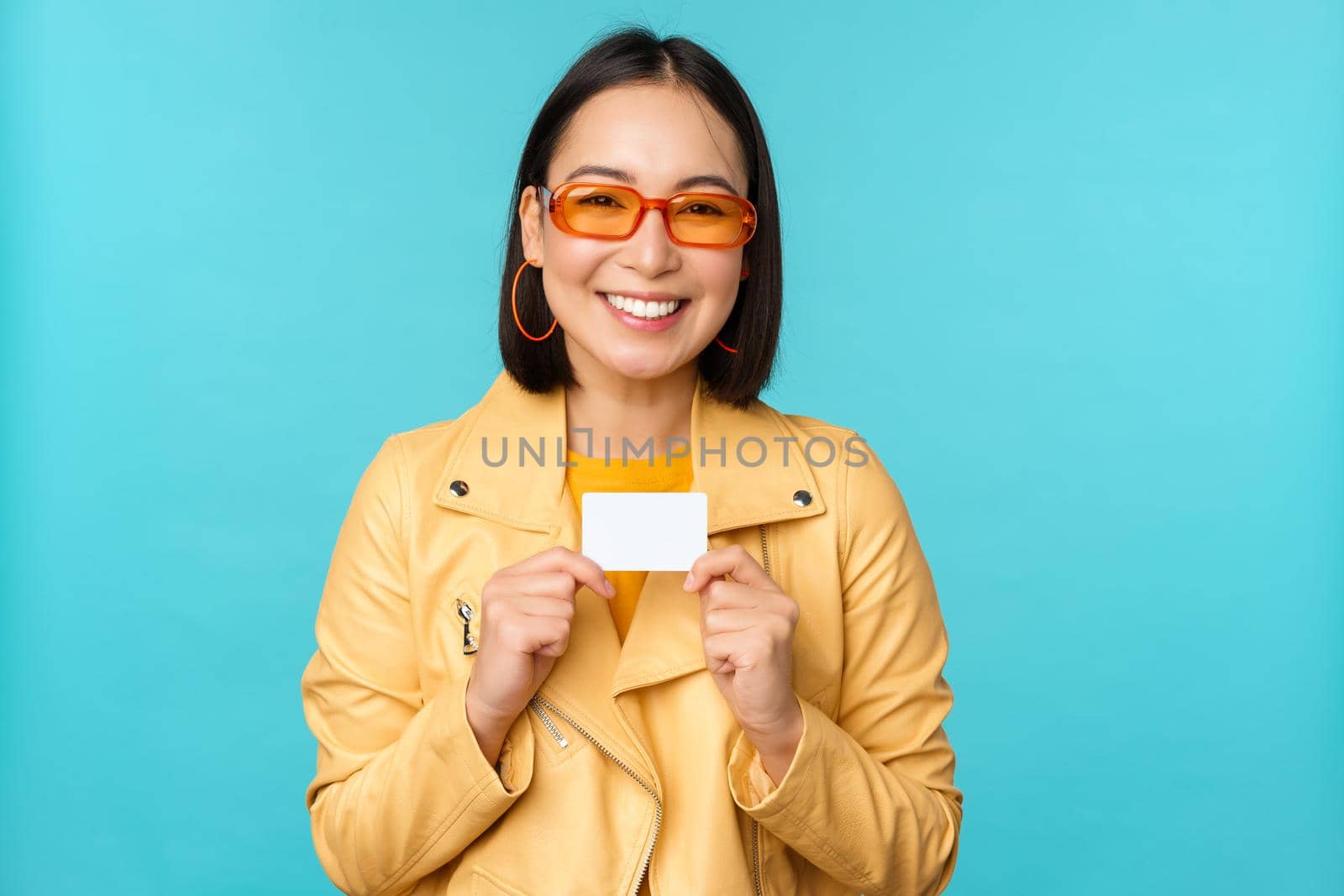 Young beautiful asian woman showing credit card, smiling, choosing bank, standing over blue background by Benzoix