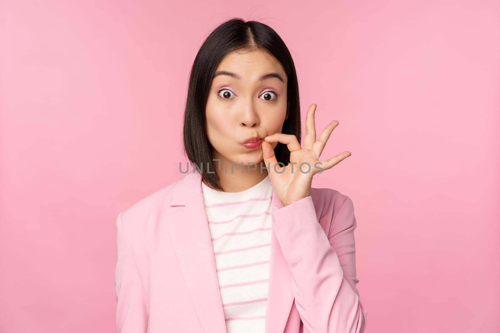 Portrait of asian corporate woman showing mouth seal, close shut lips on key gesture, promise keep secret, standing over pink background in suit.