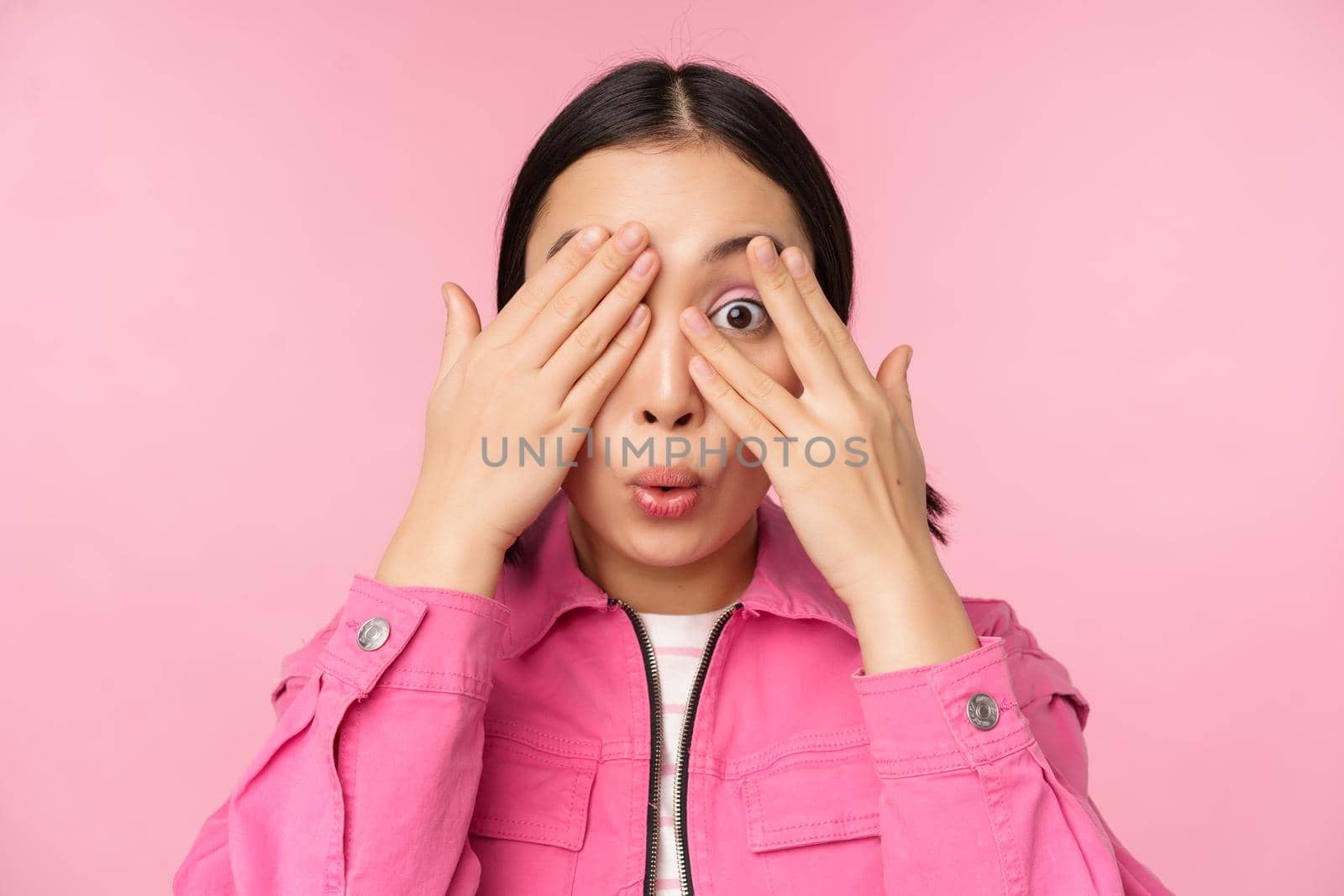 Close up portrait of young asian girl looking surprised, express amazement and wonder, peeking through fingers, standing over pink background by Benzoix