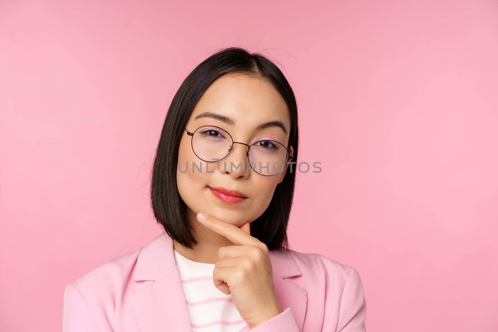 Korean businesswoman thinking, wearing glasses, looking thoughtful at camera, making decision, standing over pink background.