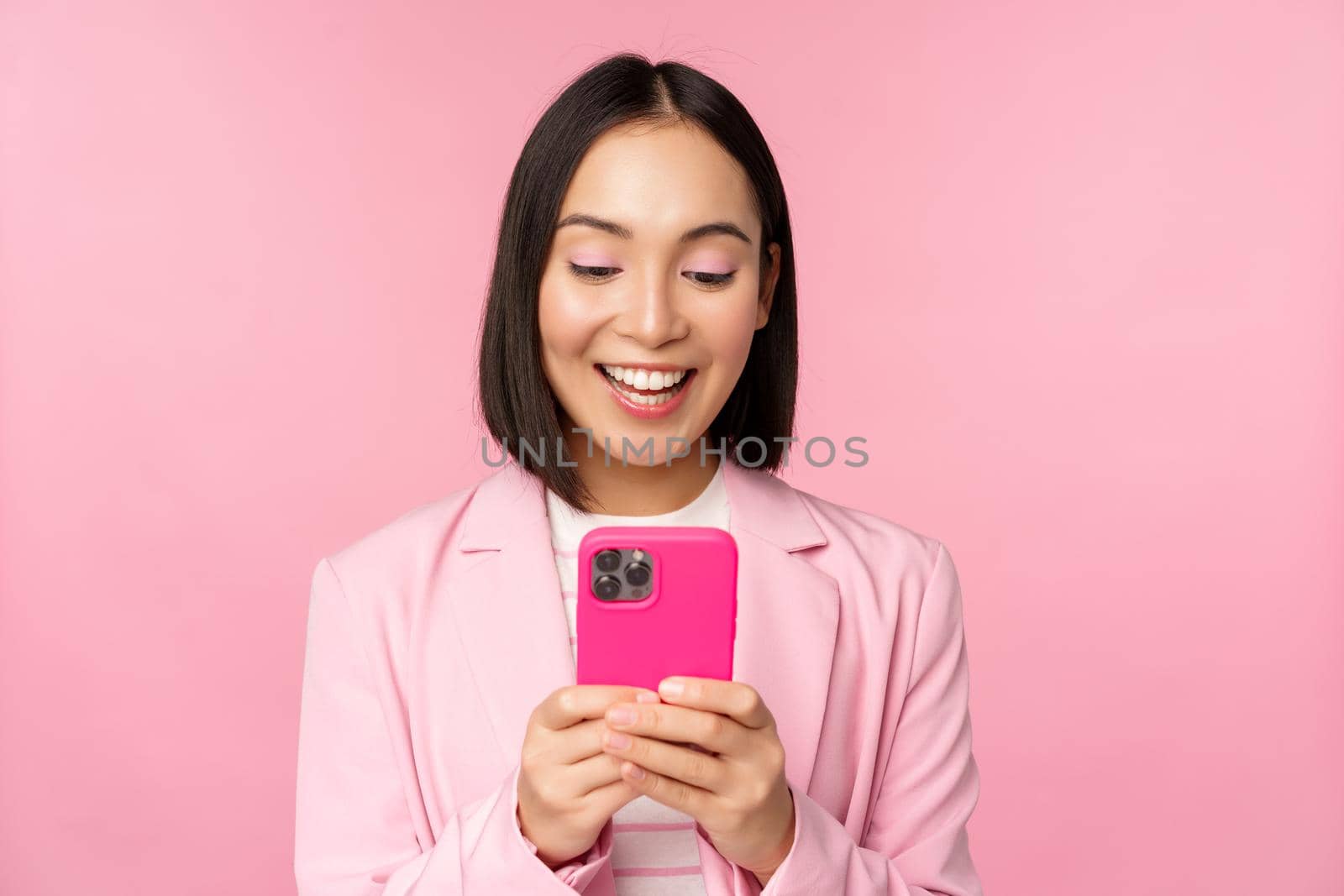 Portrait of asian girl in suit with smartphone, smiling and looking happy, standing over pink studio background by Benzoix
