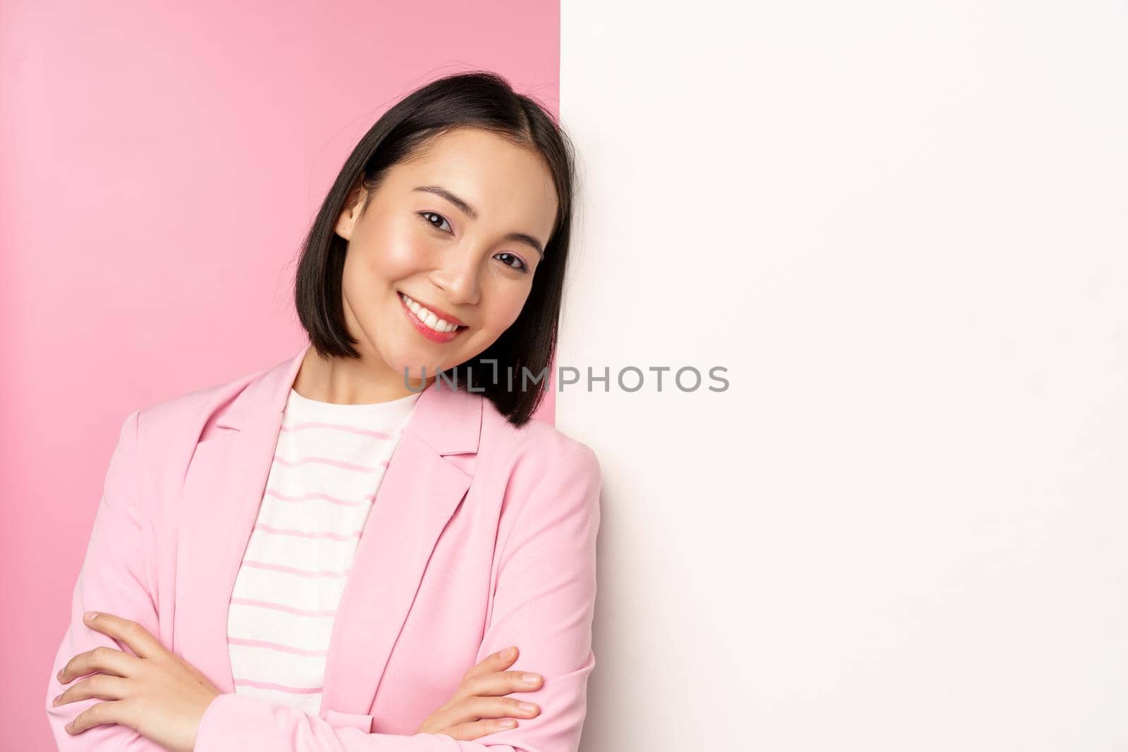 Confident, successful japanese office lady in suit, cross arms, looking as professional at camera, leaning on white wall with advertisement, empty copy space for logo, pink background.
