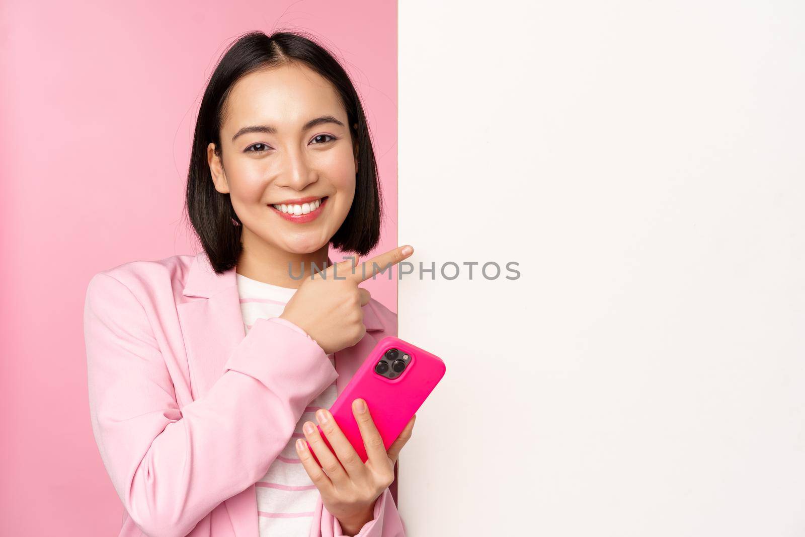 Image of smiling asian corporate woman in suit, holding smartphone, pointing at board, showing chart or information logo on empty white wall, standing over pink background.
