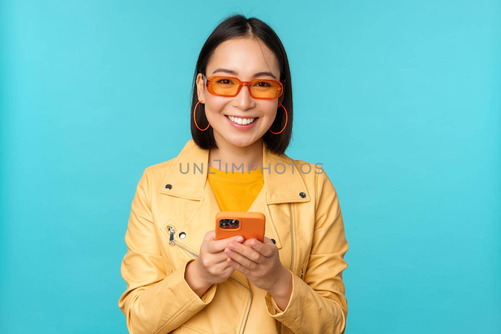 Smiling asian girl in sunglasses, using smartphone app, holding mobile phone, standing over blue background. Copy space