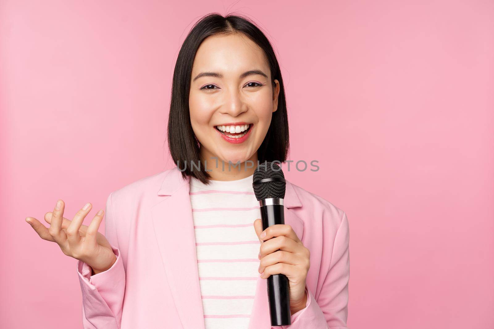 Image of enthusiastic asian businesswoman giving speech, talking with microphone, holding mic, standing in suit against pink studio background.
