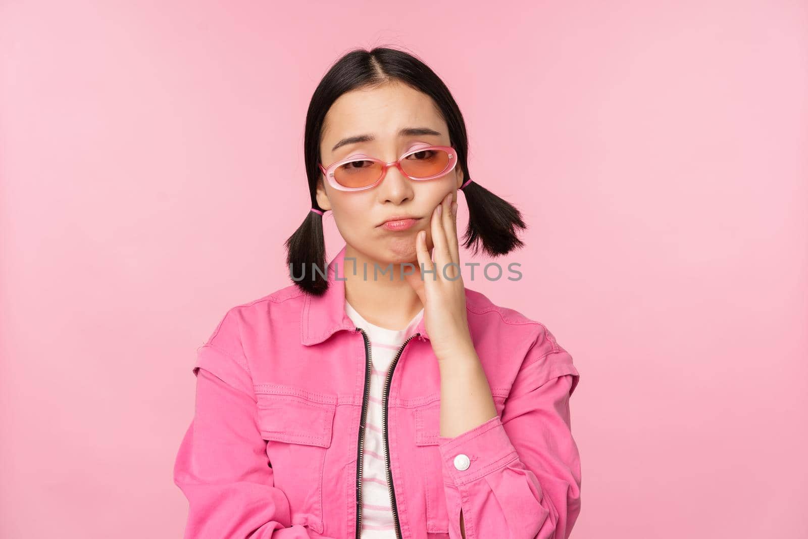 Image of sad asian girl sulking, touching her cheek, pouting disappointed, has toothache, standing over pink background.