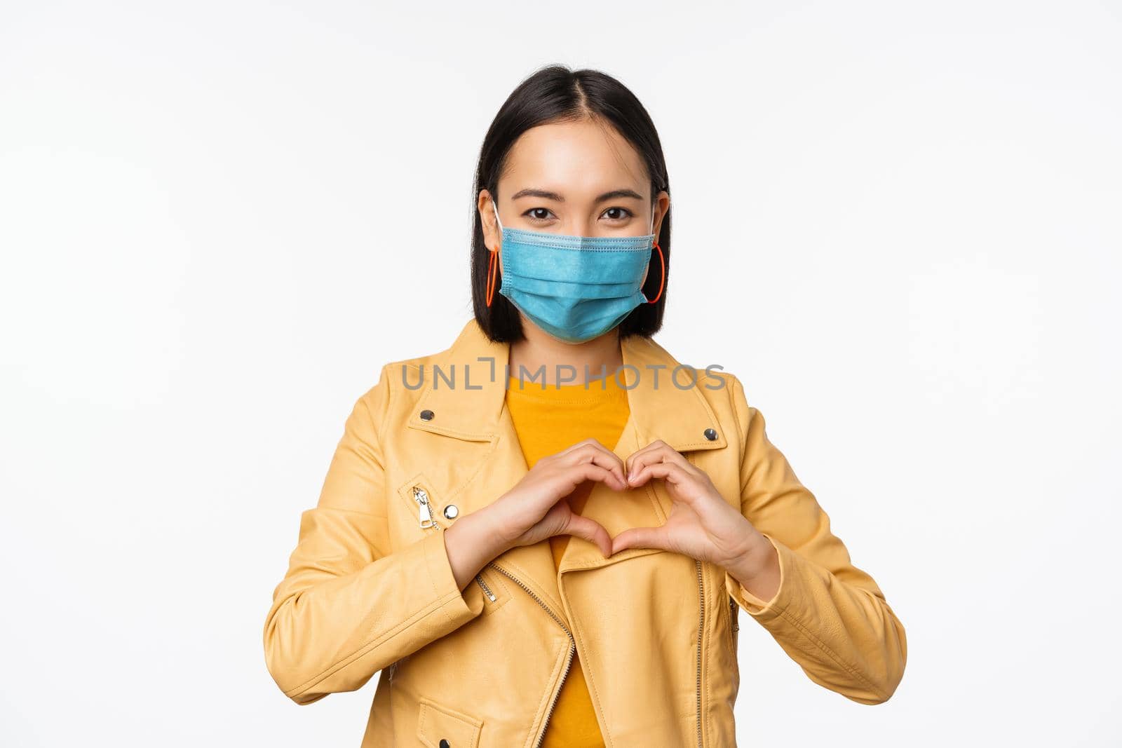 Covid-19 and people concept. Young asian woman in medical face mask, showing heart gesture and smiling with care, white background by Benzoix