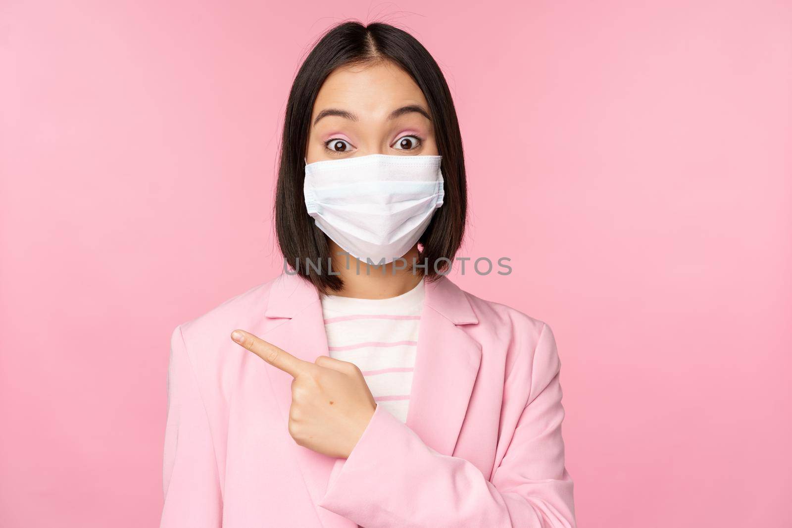 Portrait of asian businesswoman in medical face mask and suit, pointing finger left, showing advertisement, company banner, studio pink background by Benzoix