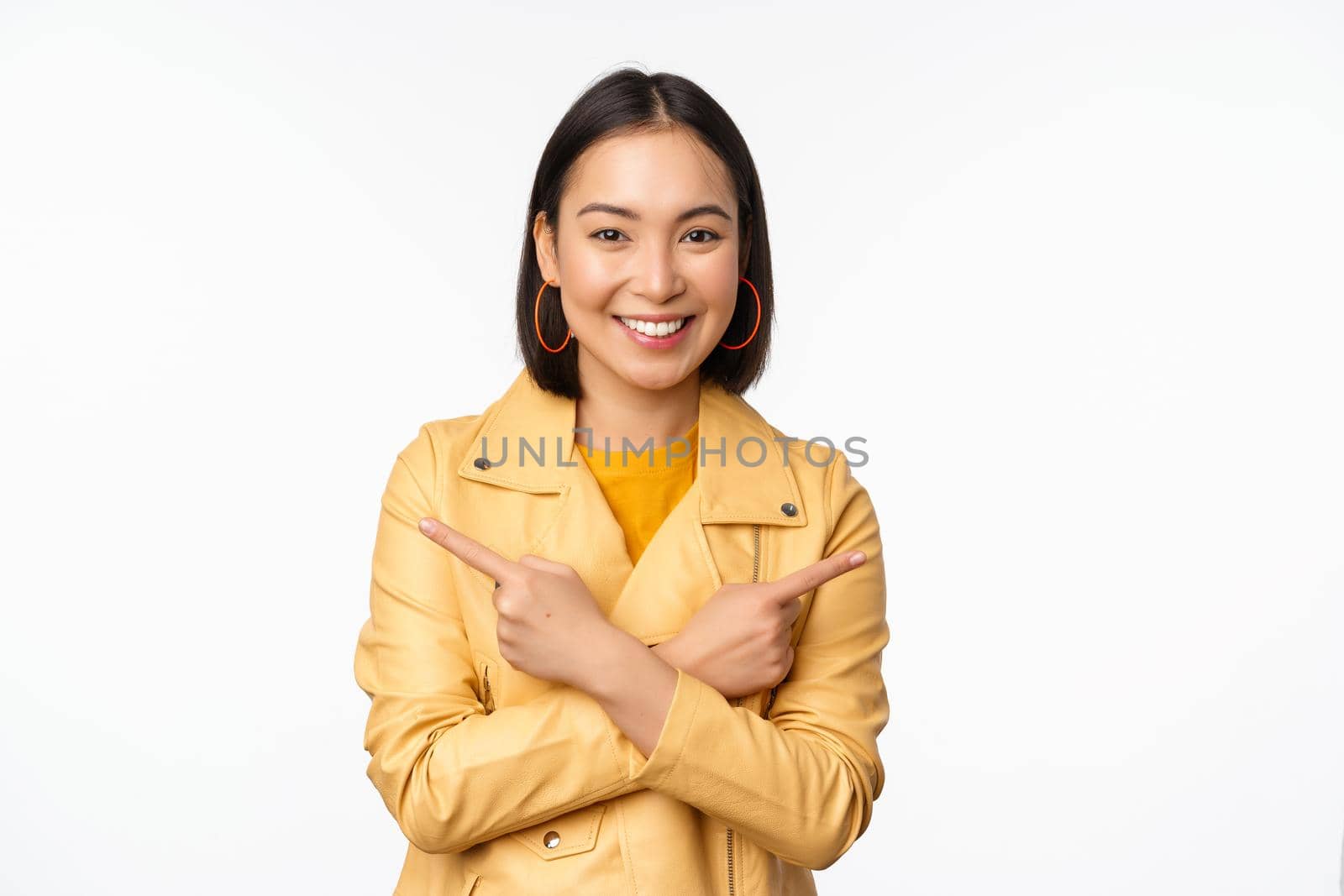 Image of indecisive asian girl, pointing fingers sideways, pointing left and right, choosing variant, deciding, standing over white background. Copy space