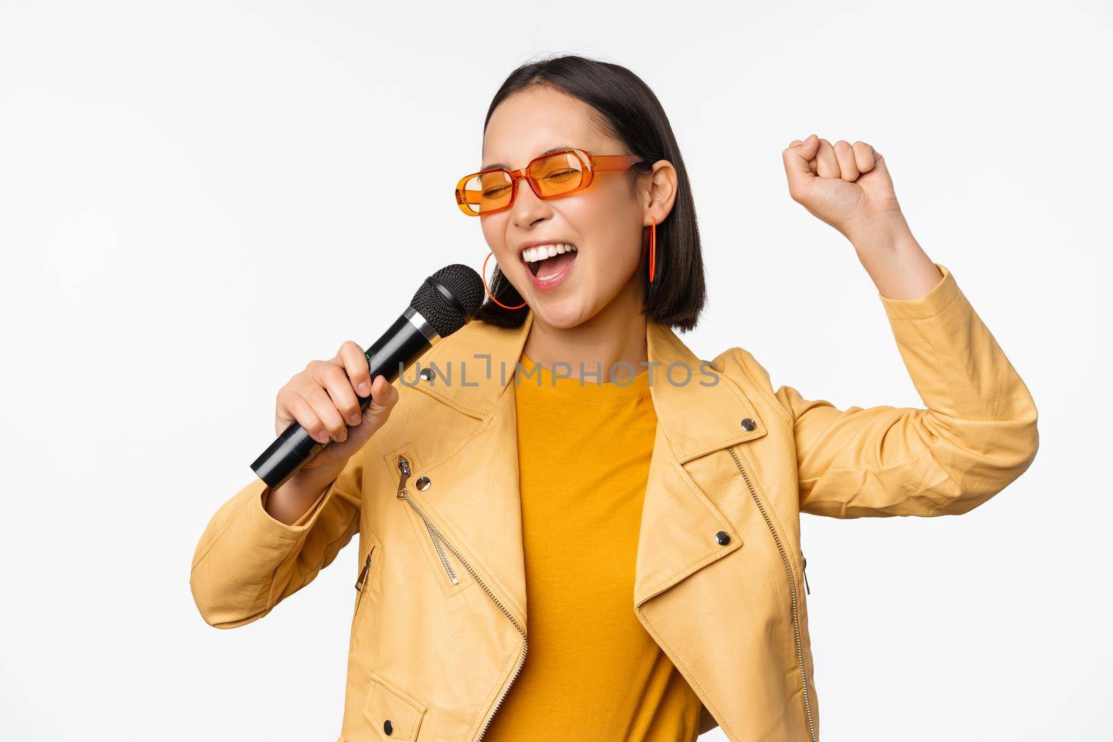 Stylish asian girl in sunglasses, singing songs with microphone, holding mic and dancing at karaoke, posing against white background by Benzoix