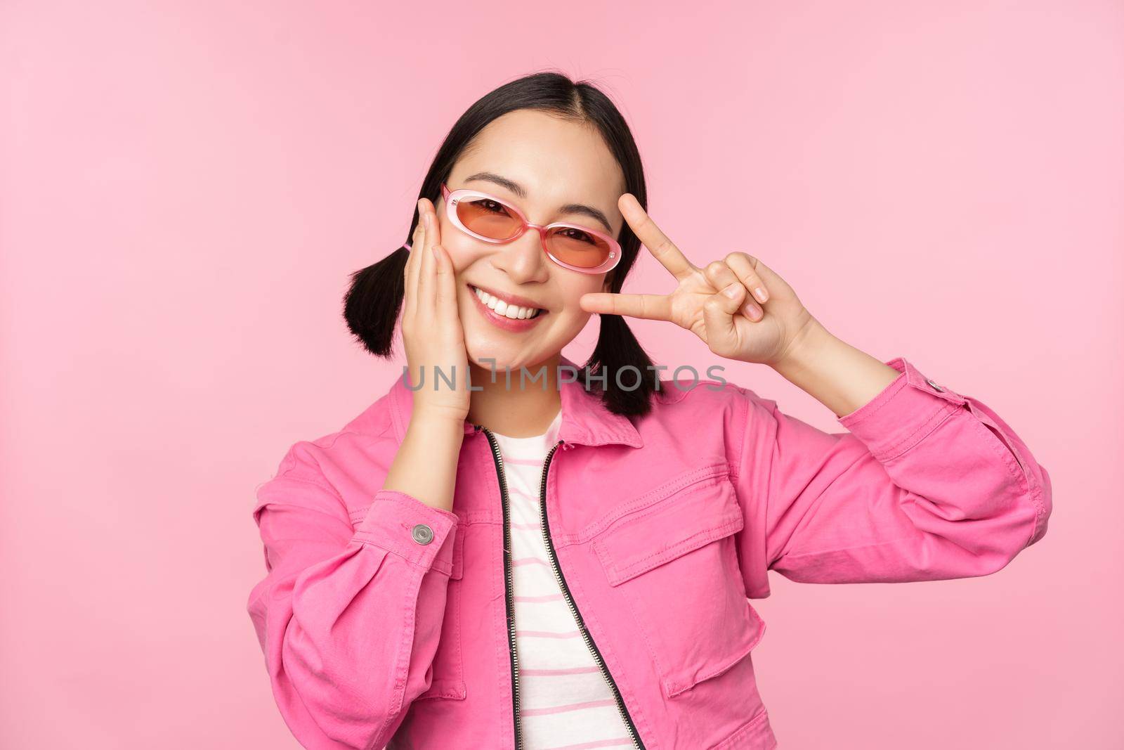 Portrait of kawaii asian girl in sunglasses, showing peace, v-sign near eye and smiles cute at camera, posing in trendy clothes against pink background.