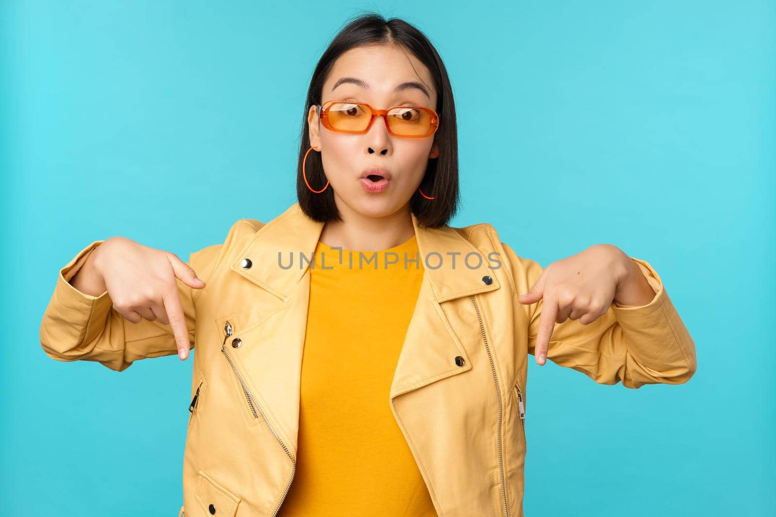 Portrait of stylish asian girl, wears sunglasses, smiles and points fingers down, shows advertisement, stands over blue background by Benzoix