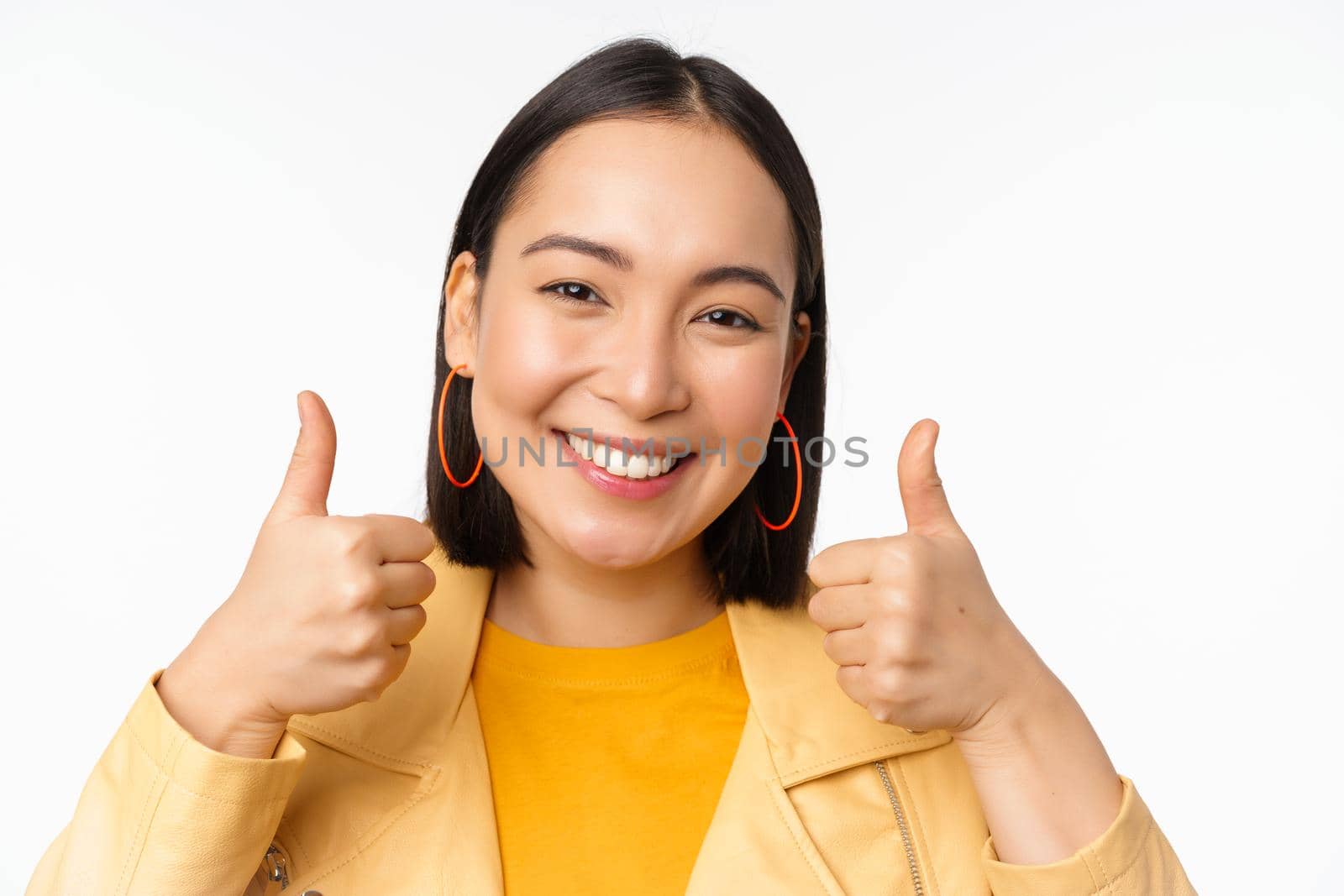 Smiling asian girl showing thumbs up, looking pleased, approve smth, standing over white background by Benzoix