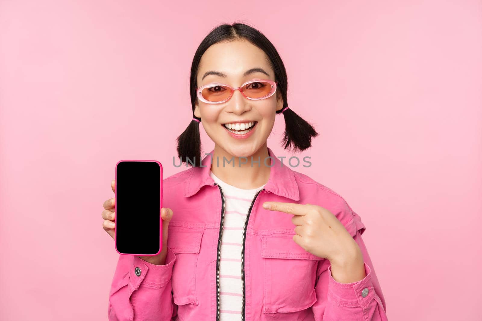 Enthusiastic asian woman in stylish clothes, sunglasses, pointing finger at mobile phone screen, showing smartphone application, standing over pink background by Benzoix