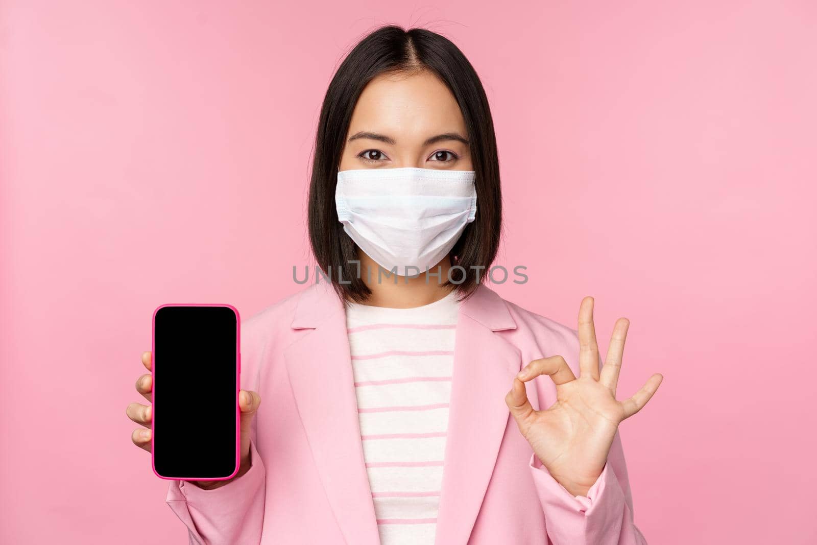 Portrait of asian businesswoman in medical face mask covid-19, showing smartphone screen and okay sign, pink background.