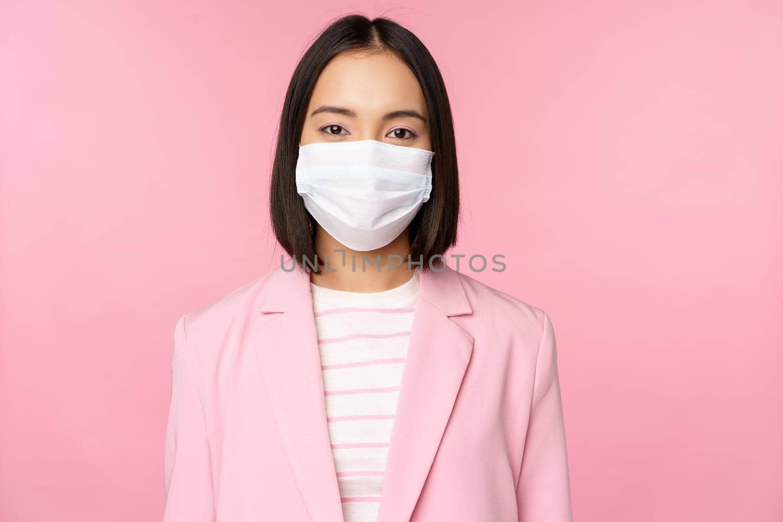 Portrait of asian businesswoman in medical face mask, wearing suit, concept of office work during covid-19 pandemic, standing over pink background.