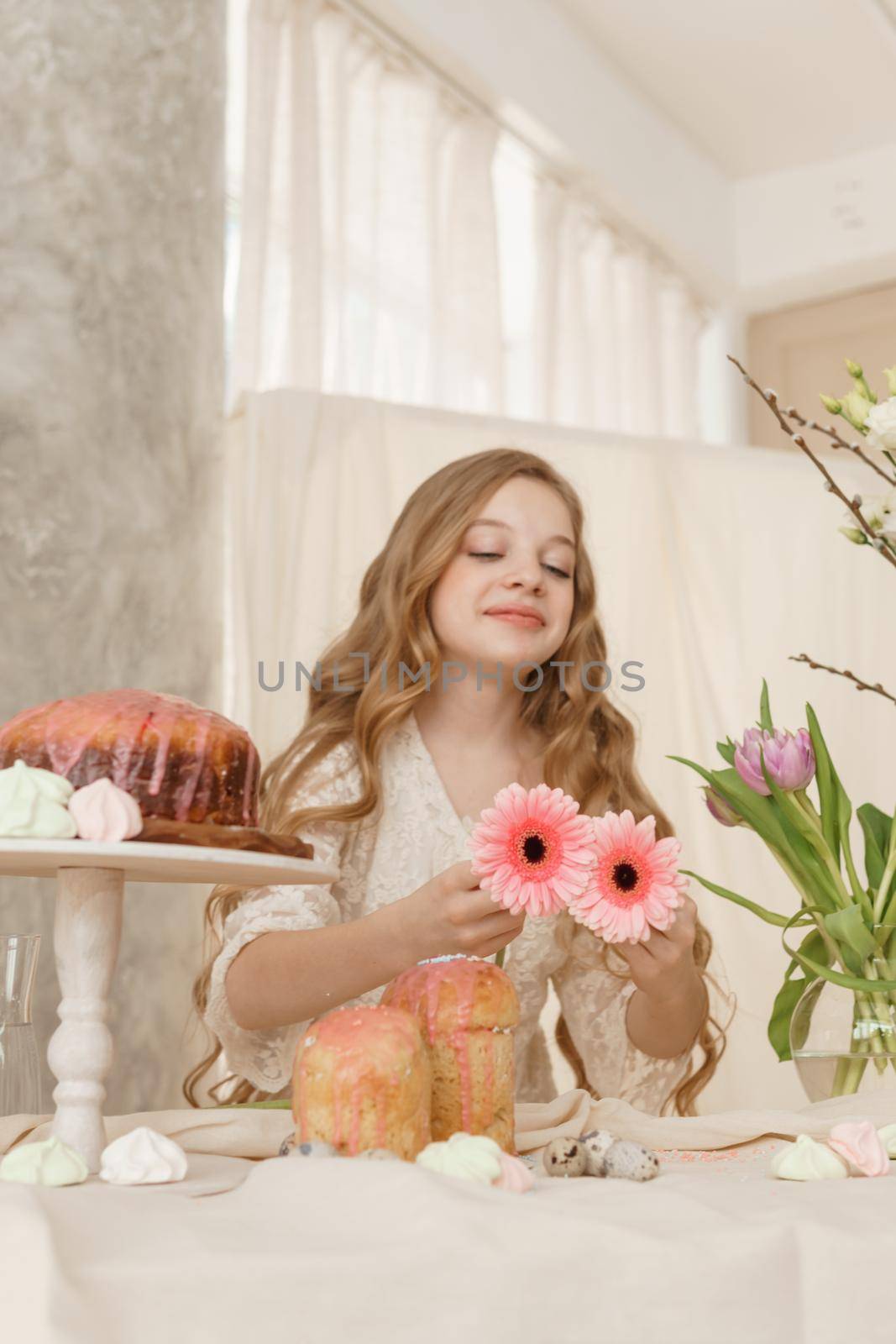 A girl with long hair in a light dress is sitting at the Easter table with cakes, spring flowers and quail eggs. Happy Easter celebration