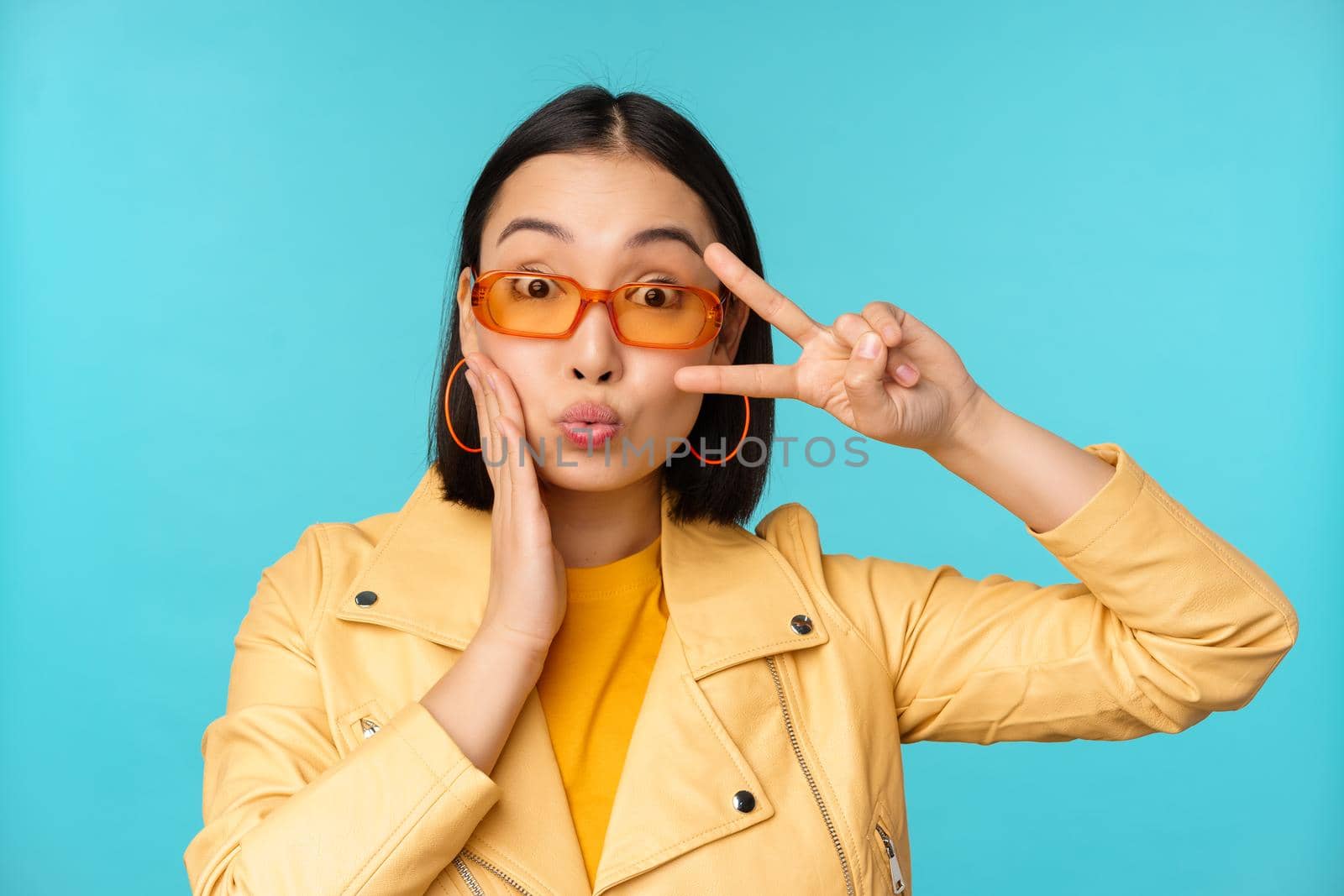 Portrait of attractive korean woman in sunglasses, showing peace v-sign near eyes, pucker lips, kissing gesture, standing over blue background.