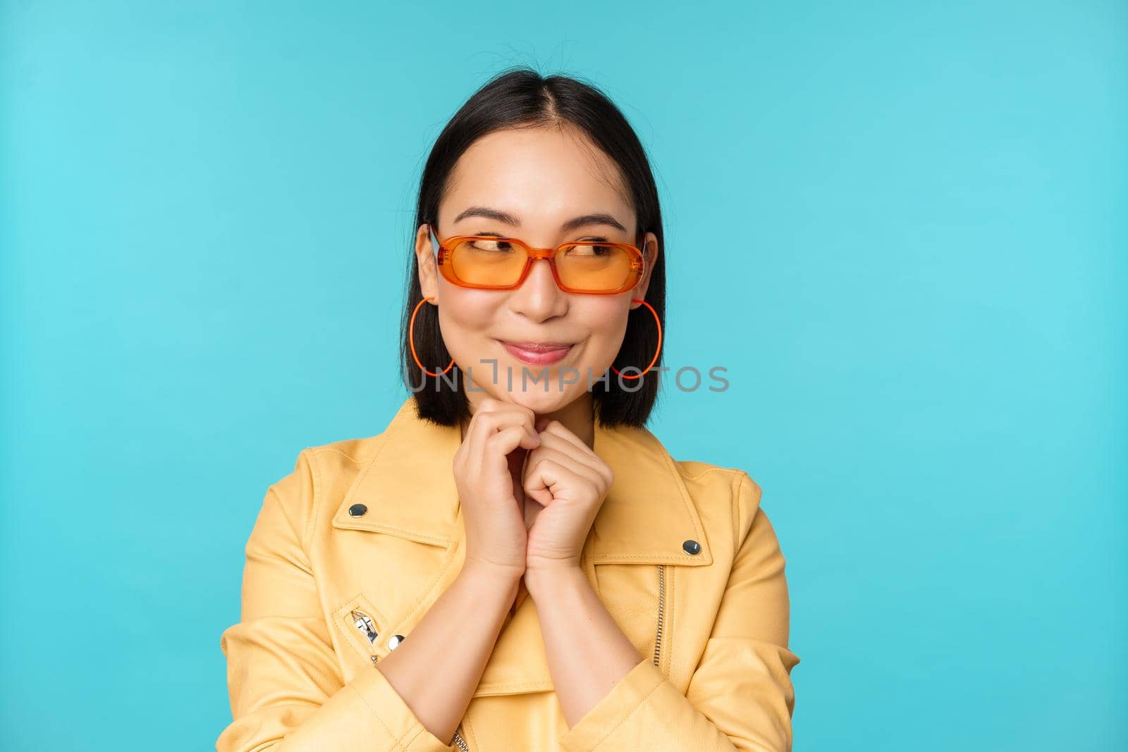 Portrait of stylish korean woman in sunglasses, smiling and looking aside at logo coquettish, standing over blue background. Copy space