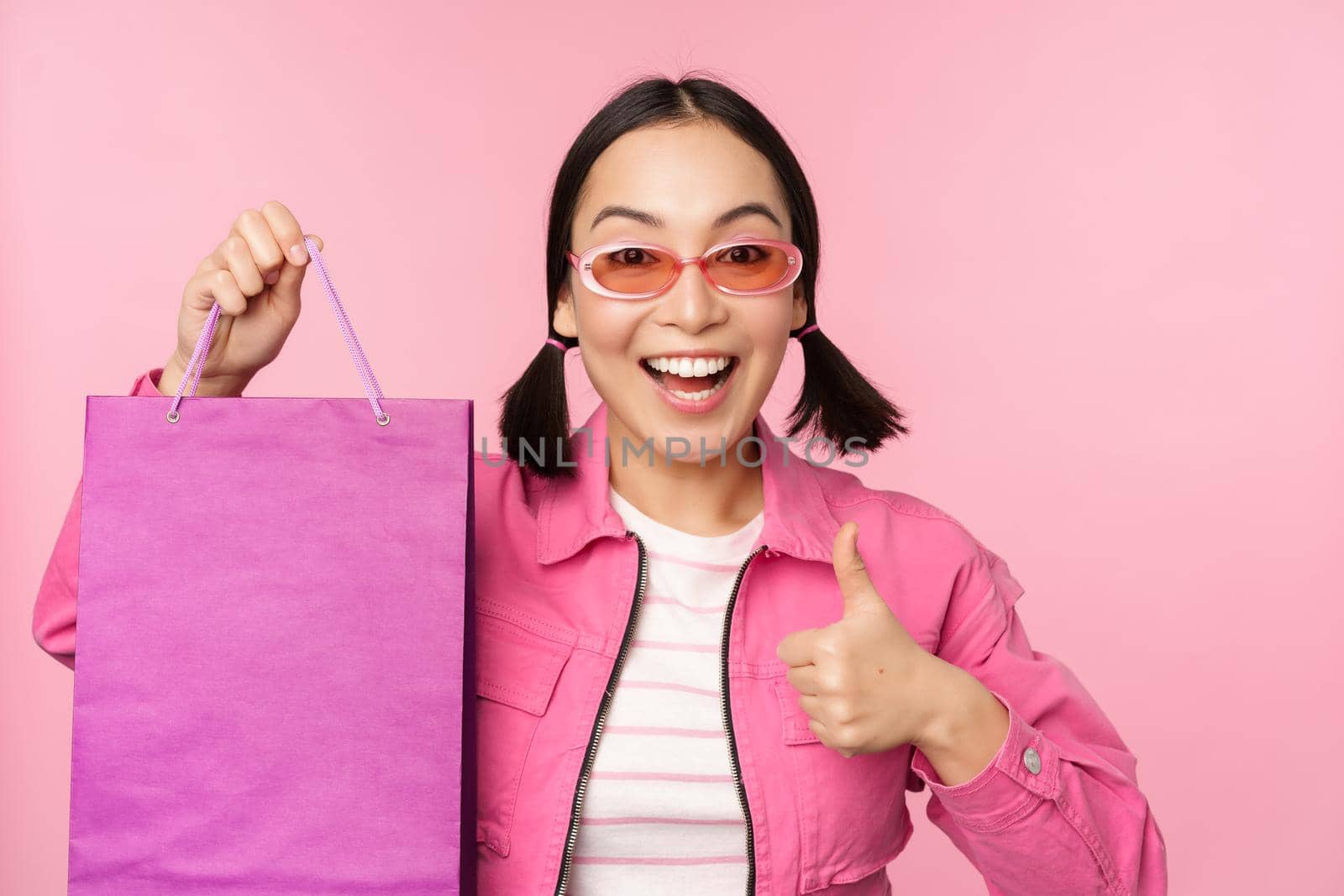 Shopping. Stylish asian girl in sunglasses, showing bag from shop and smiling, recommending sale promo in store, standing over pink background.