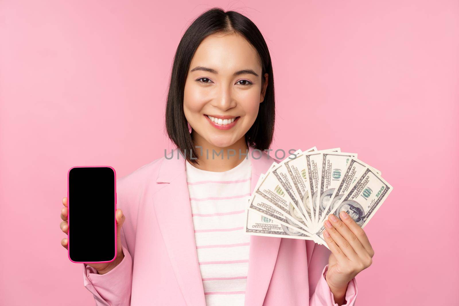 Image of korean successful corporate woman showing money, dollars and smartphone app screen, interface of mobile phone application, concept of investment and finance, pink background by Benzoix