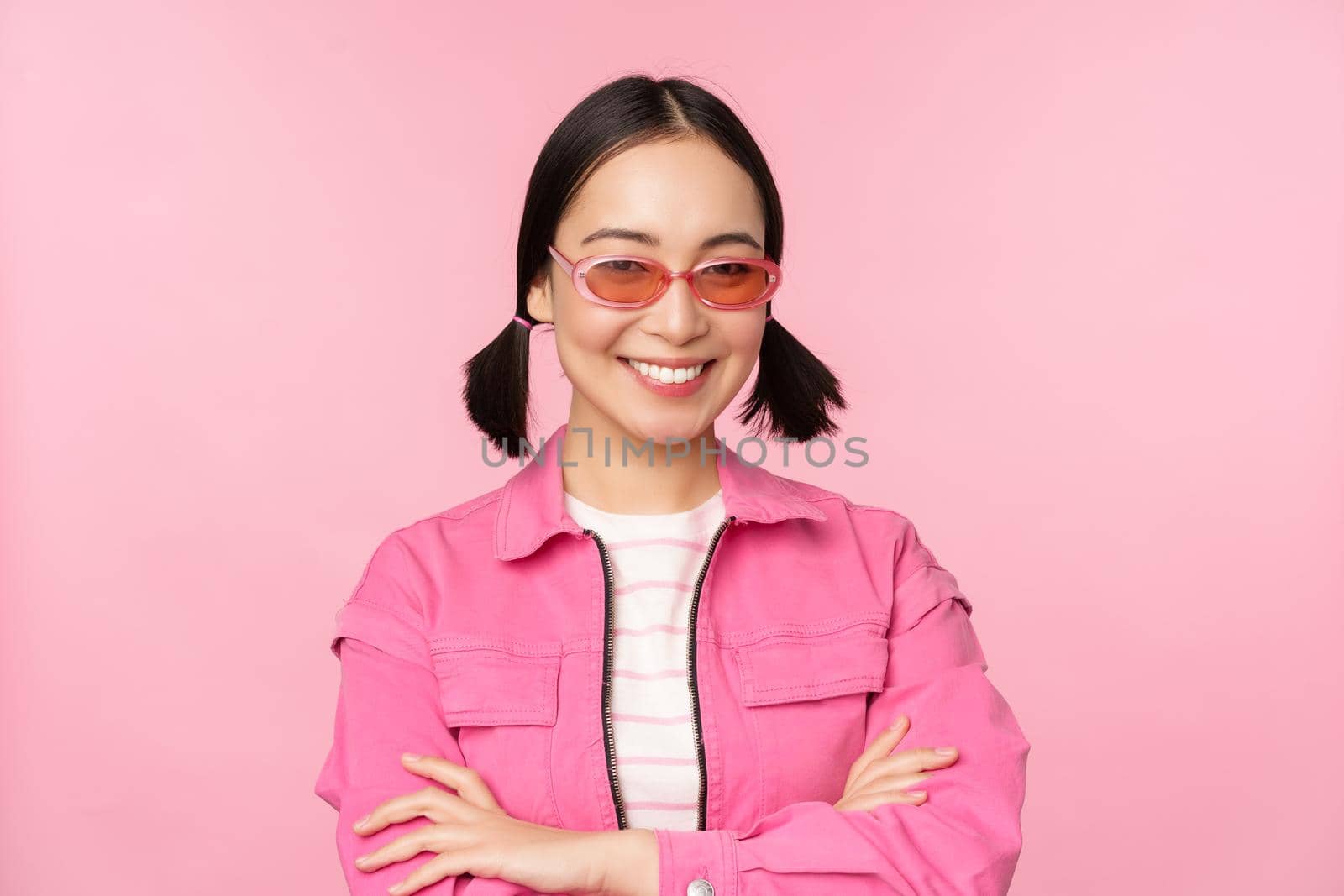 Stylish asian girl in pink clothes and sunglasses, smiling and looking happy at camera, standing over studio background by Benzoix