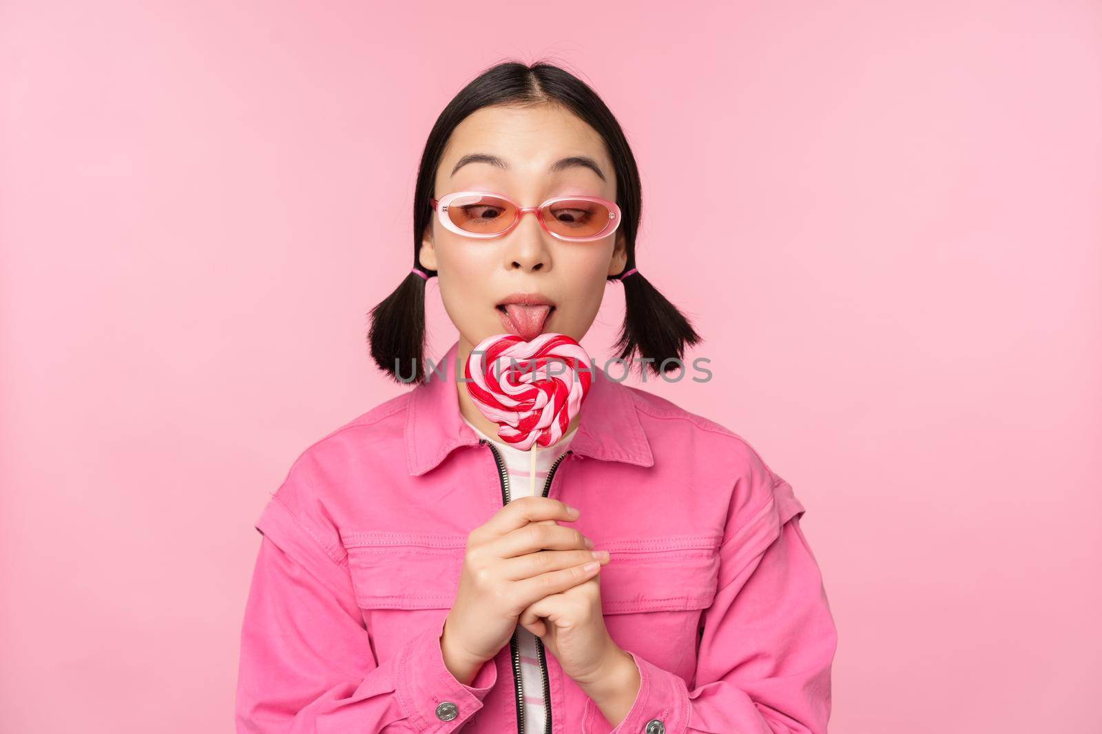 Silly and cute asian female model licking lolipop, eating candy sweet and smiling, looking excited, standing over pink background by Benzoix