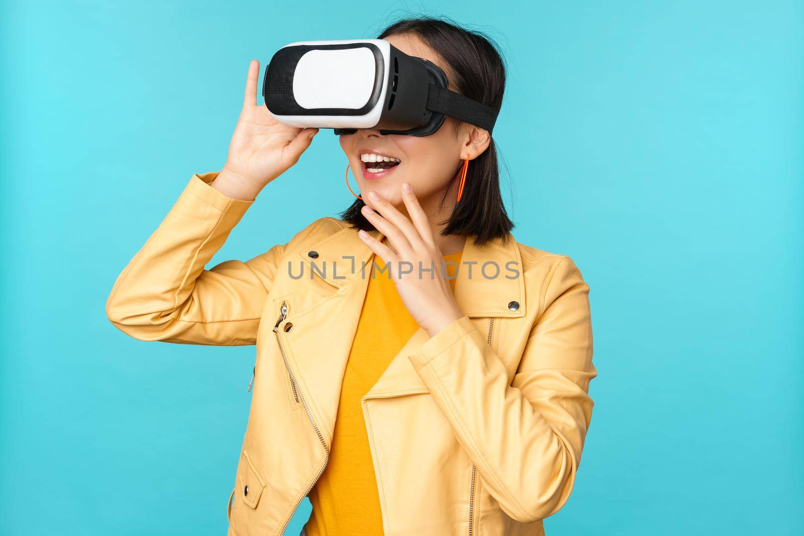 Portrait of happy asian female model using VR headset, smiling and laughing in virtual reality glasses, standing over blue background.