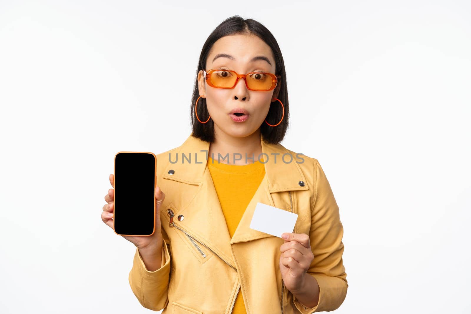 Online shopping and people concept. Stylish asian woman showing mobile phone screen and credit card, smartphone application, standing over white background by Benzoix