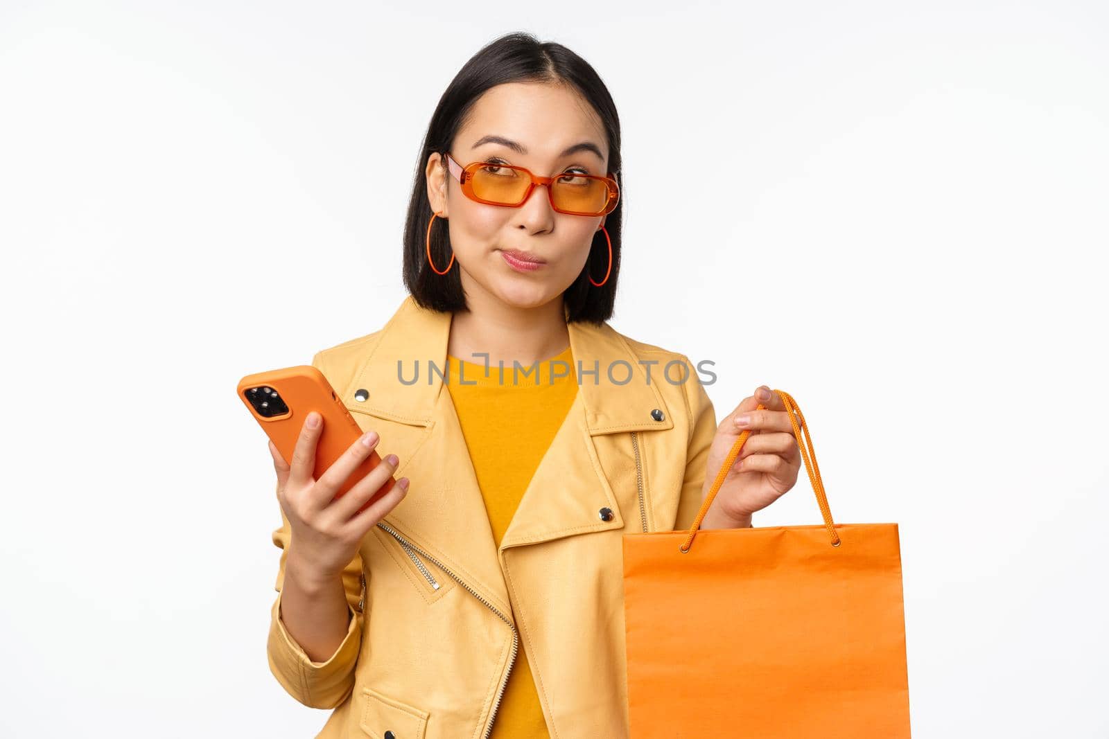 Stylish asian girl shopper, wears sunglasses, holding shopping bag and smartphone, going for discounts in stores, standing over white background.