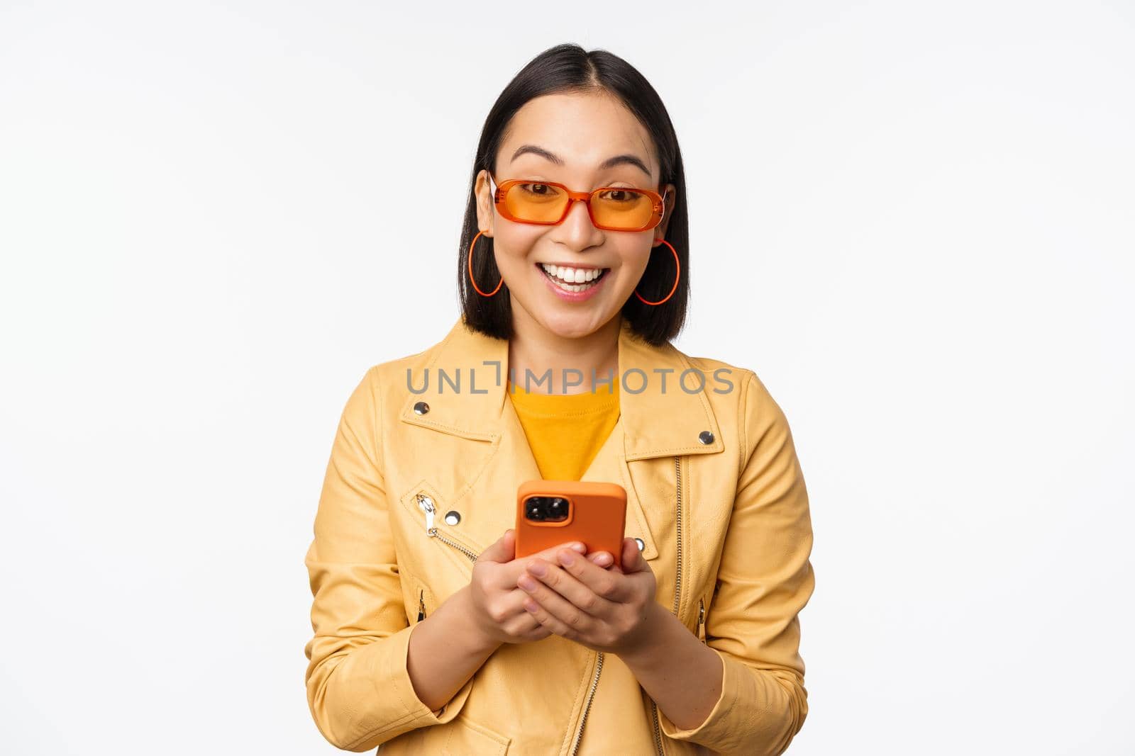 Modern asian girl in sunglasses using her mobile phone, smiling and looking happy, posing against white background. Copy space