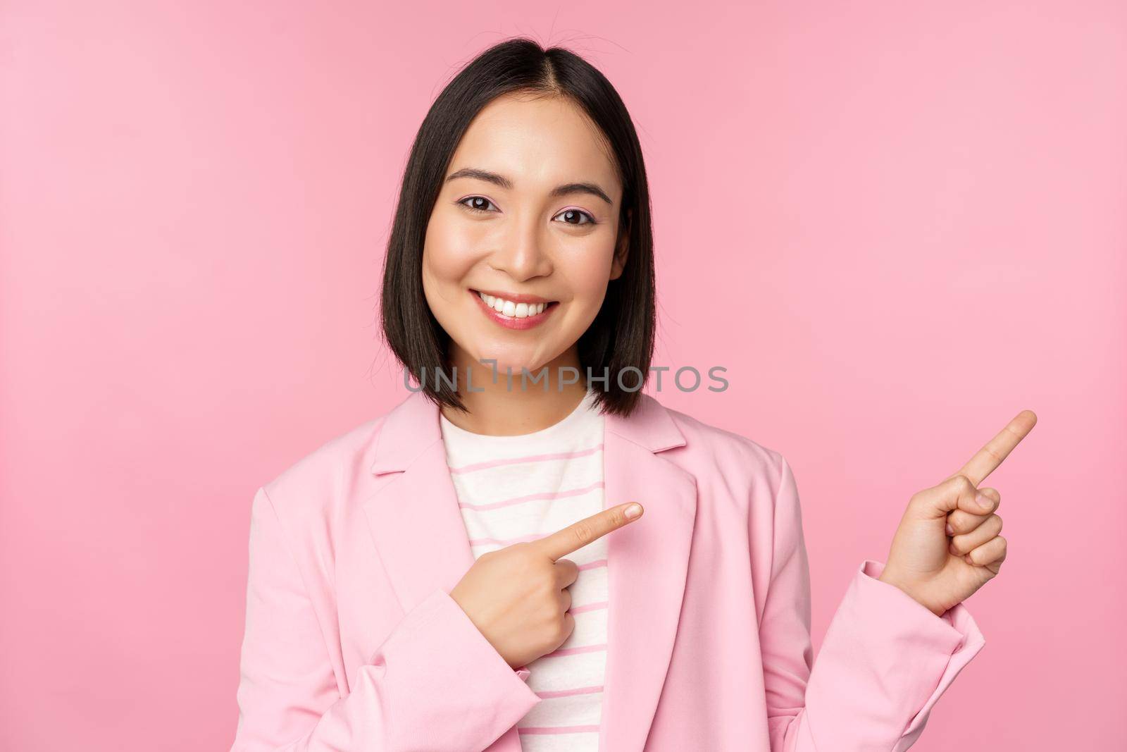 Enthusiastic professional businesswoman, saleswoman pointing fingers right, showing advertisement or company logo aside, posing over pink background.