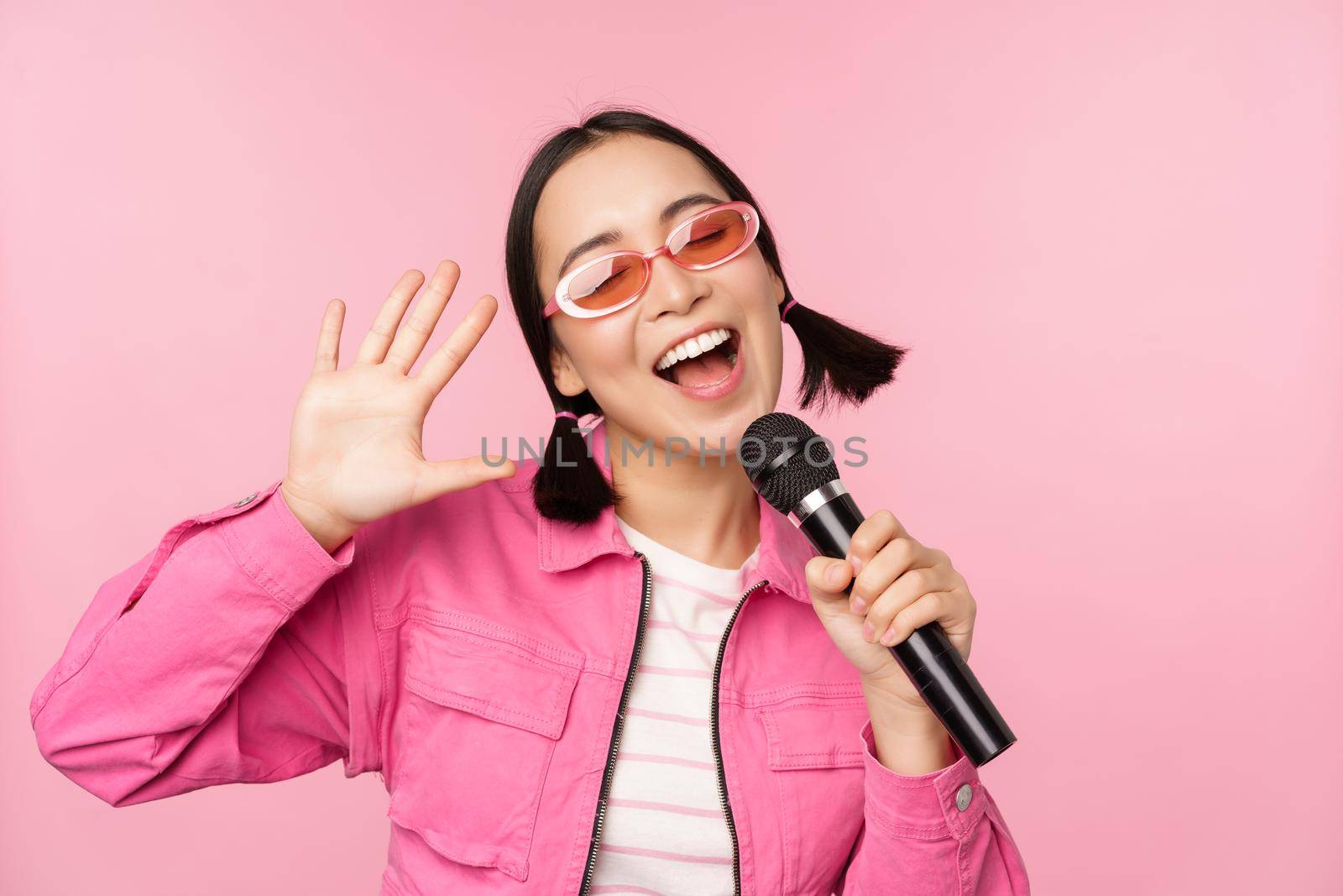 Happy beautiful asian girl singing with mic, using microphone, enjoying karaoke, posing against pink studio background by Benzoix