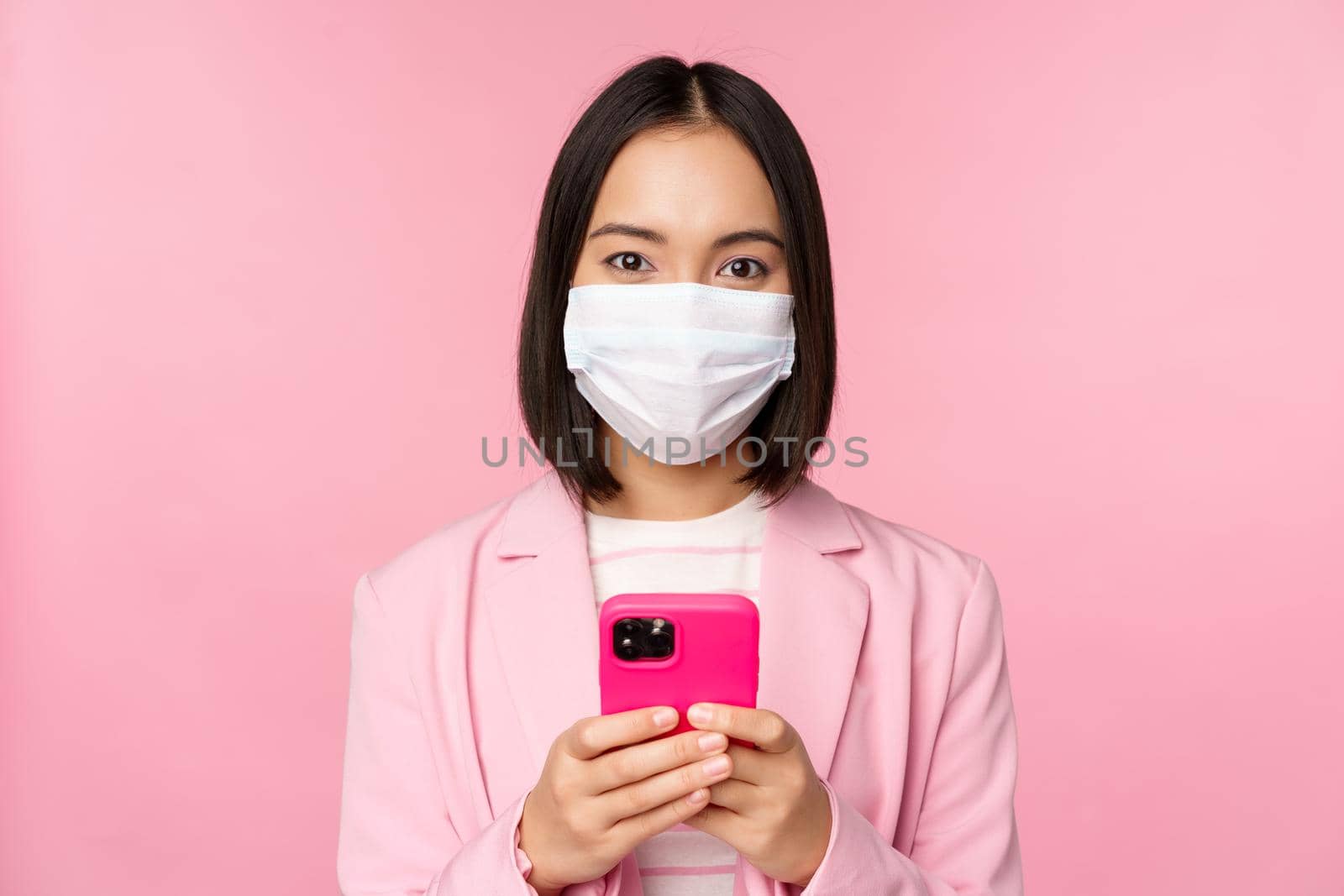 Business people and covid-19 concept. Japanese corporate office lady in suit and medical face mask, using mobile phone and smiling at camera, pink background.