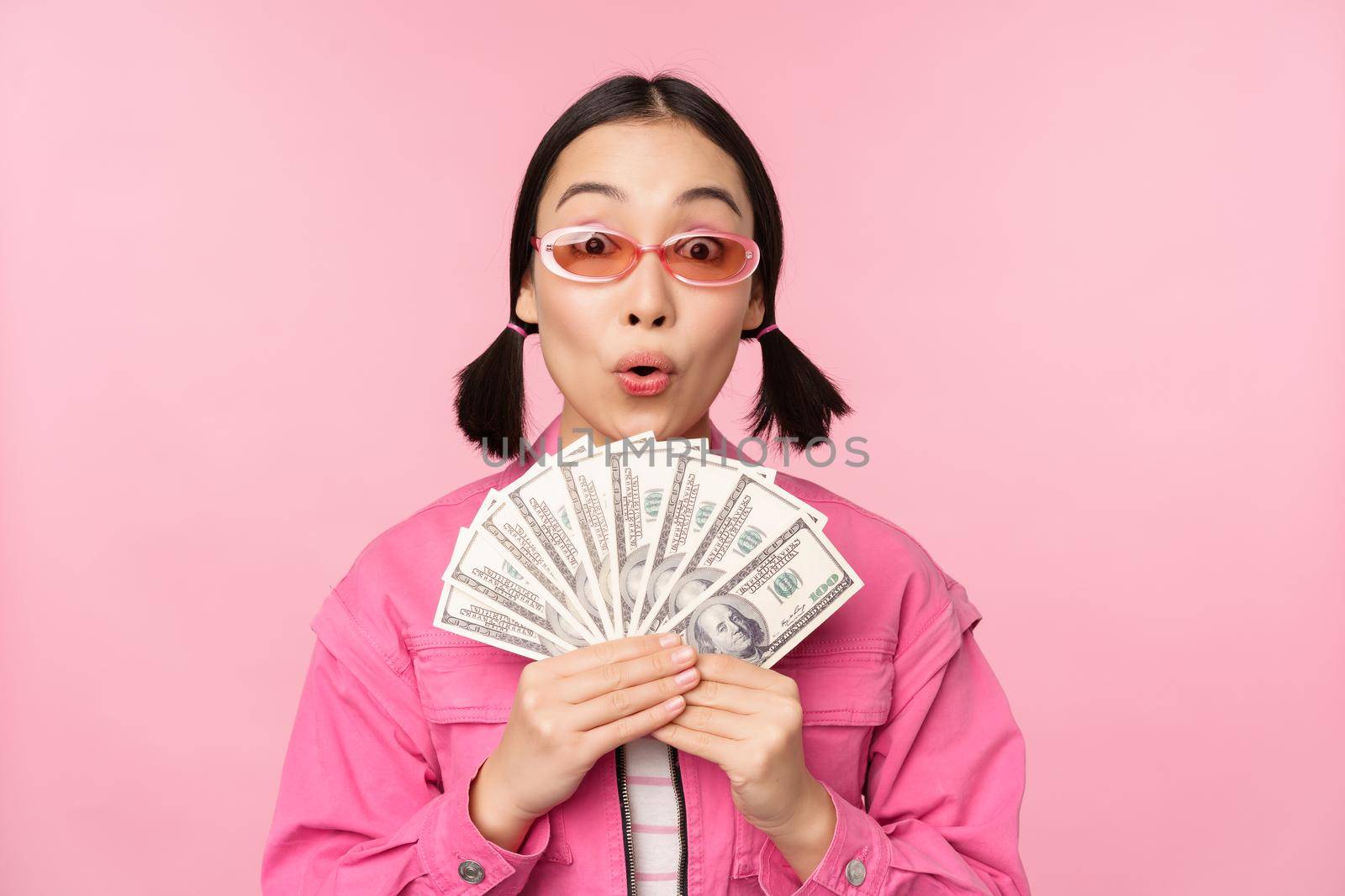 Microcredit and fast loans concept. Excited stylish korean girl, showing money, cash dollars and looking happy, standing in sunglasses over pink background by Benzoix