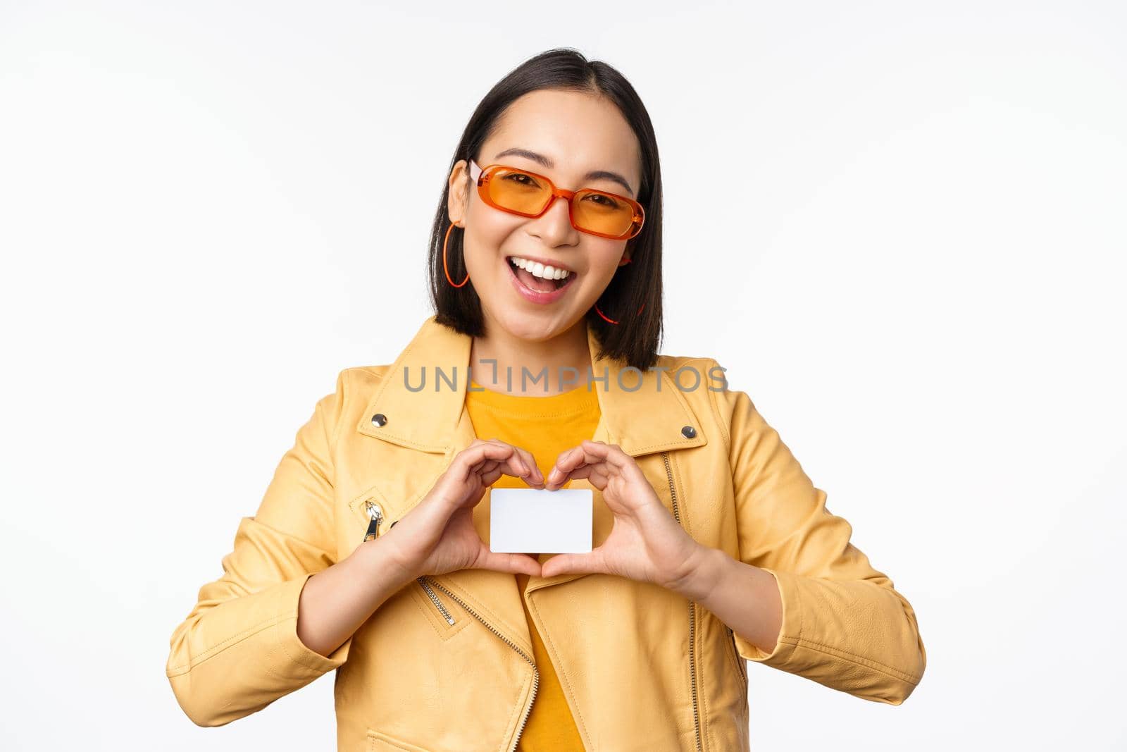 Stylish attractive asian girl in sunglasses, showing credit card and smiling, standing happy against white studio background. Copy space