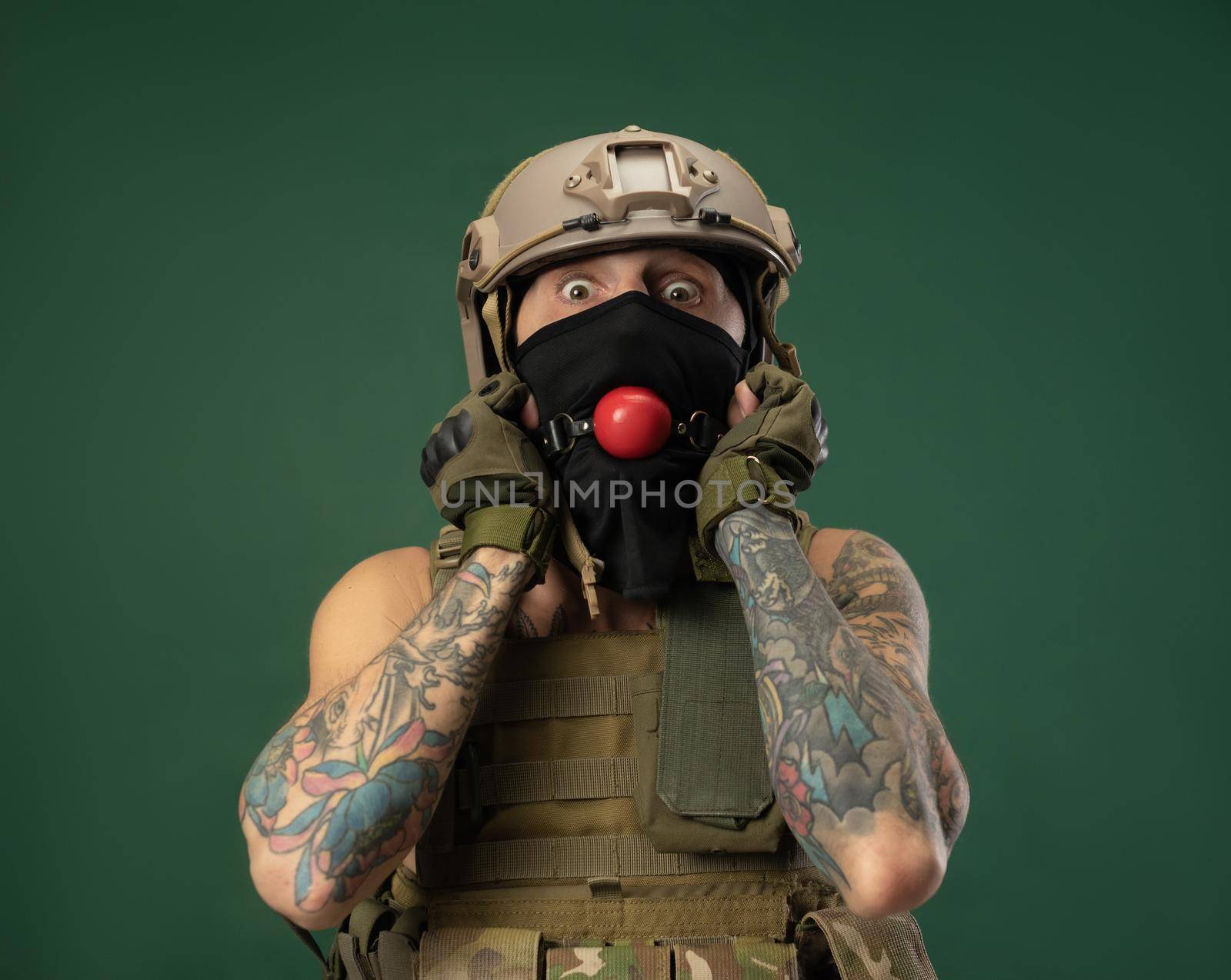 a soldier man in military clothes helmet with a bdsm gag in his mouth expresses emotions, photo joke by Rotozey