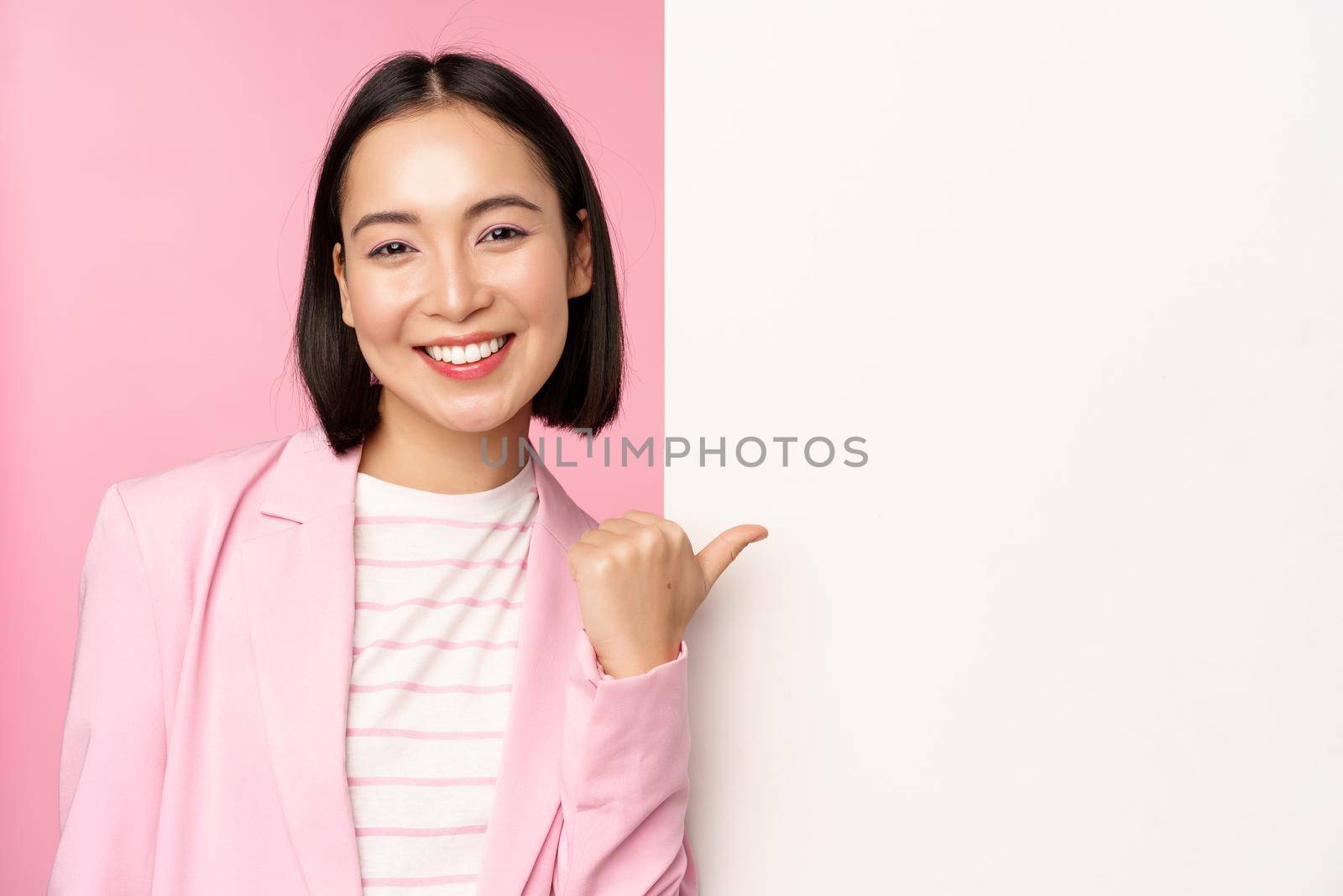 Portrait of smiling asian businesswoman in suit, corporate lady pointing finger at white empty wall, board with info or advertisement, standing over pink background.