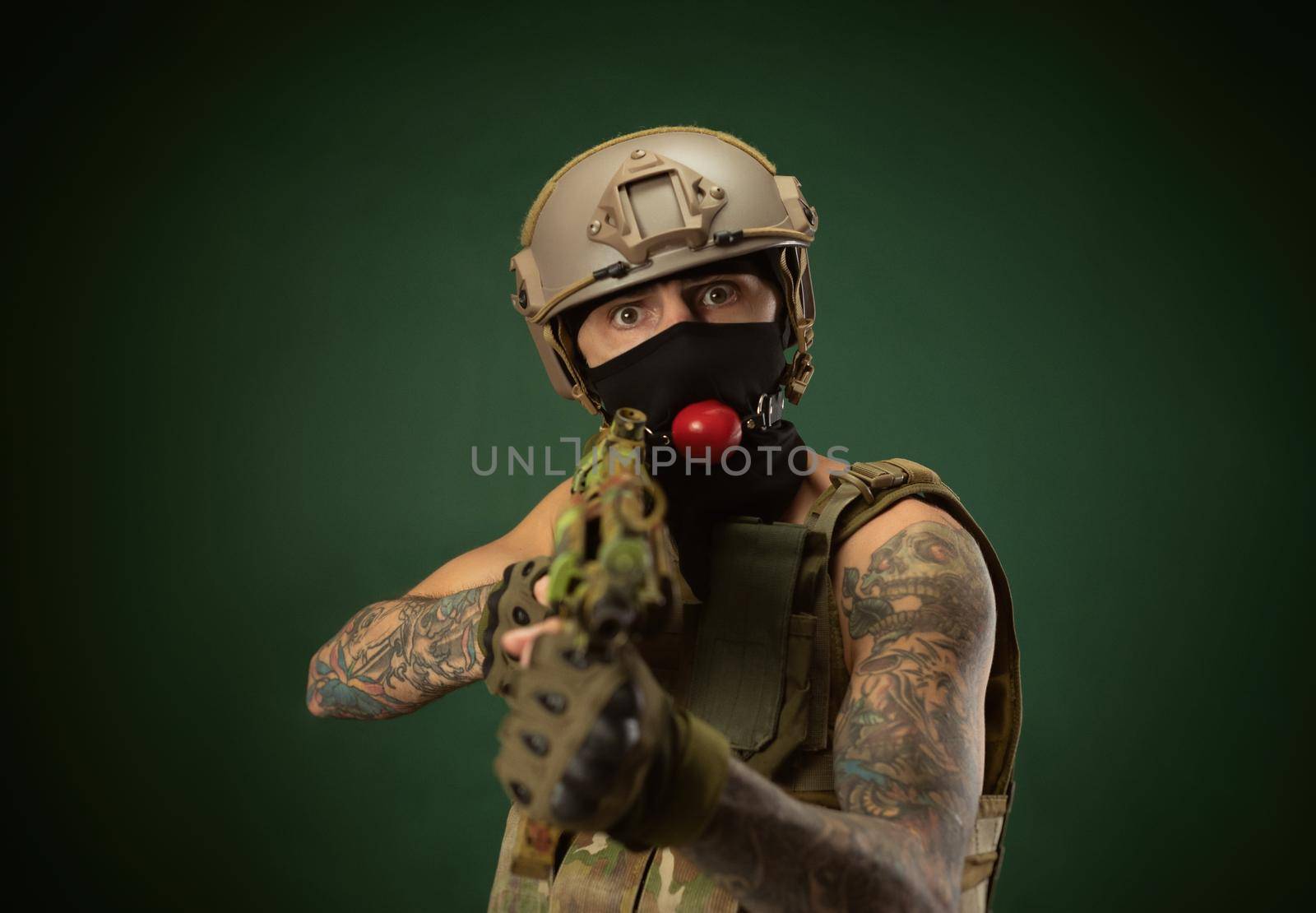 male soldier in military clothes, a helmet with a bdsm gag in his mouth expresses emotions holding an automatic rifle in his hands, photo joke