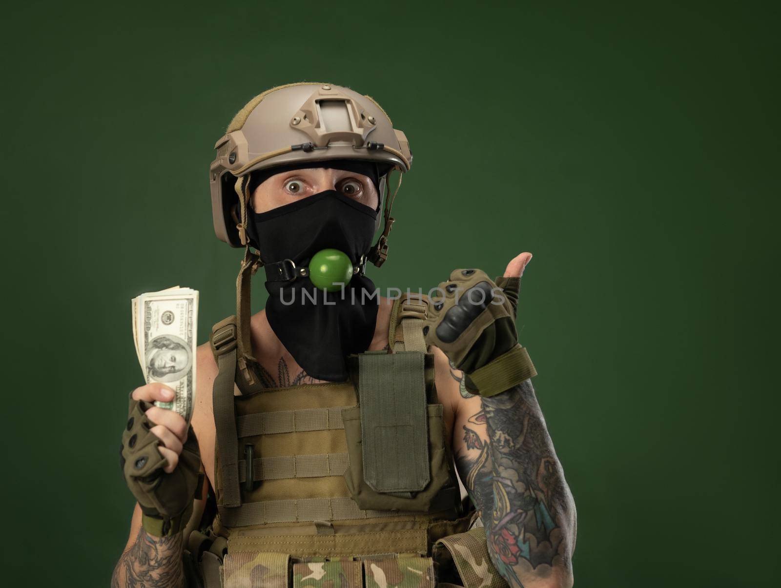 a soldier man in military clothes helmet with a bdsm gag in his mouth expresses emotions holding money dollars in his hands, photo joke by Rotozey