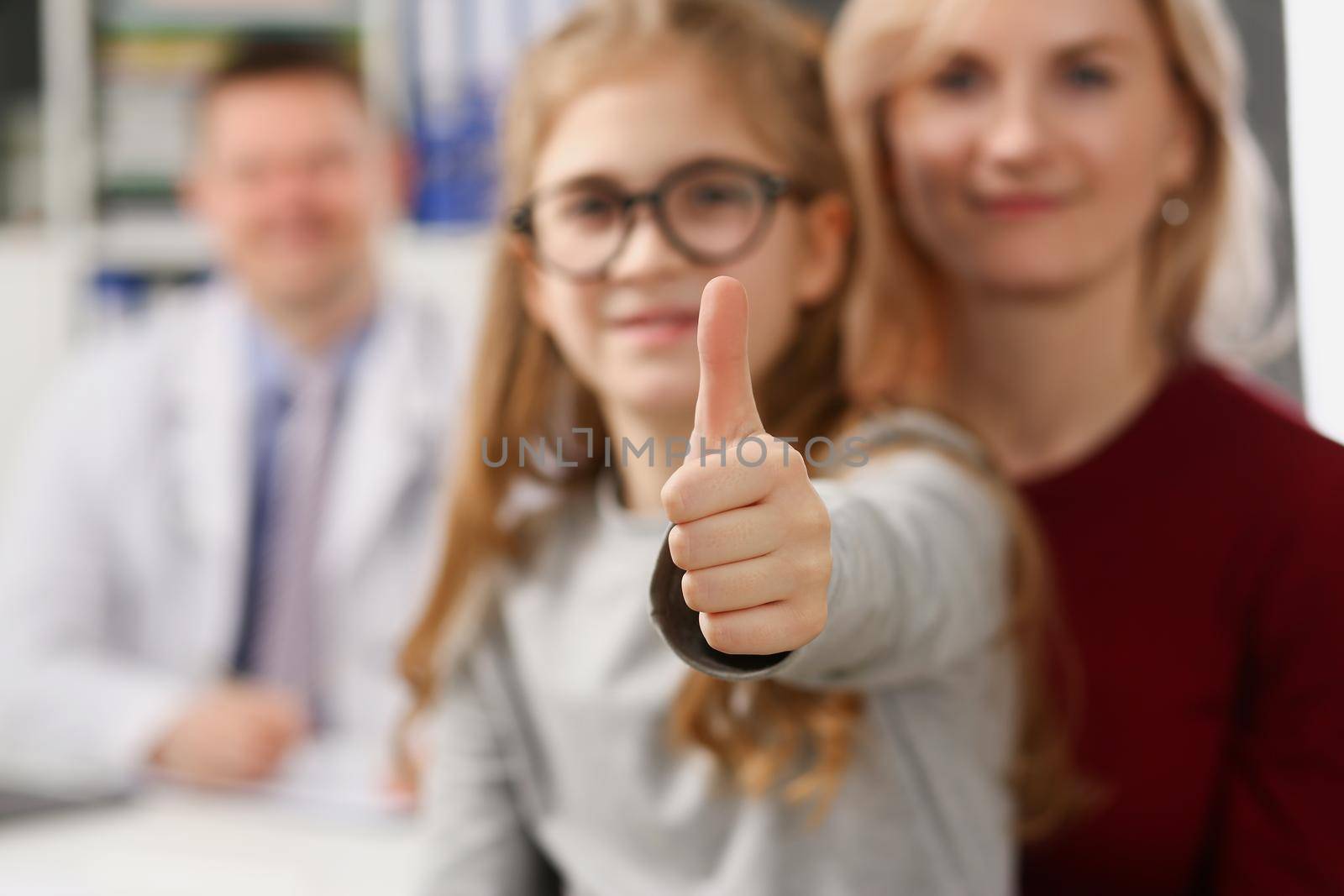 Portrait of mother and daughter on appointment in medical center, doctor smiling on background. Kid show thumbs up. Family doctor, medicine, help concept