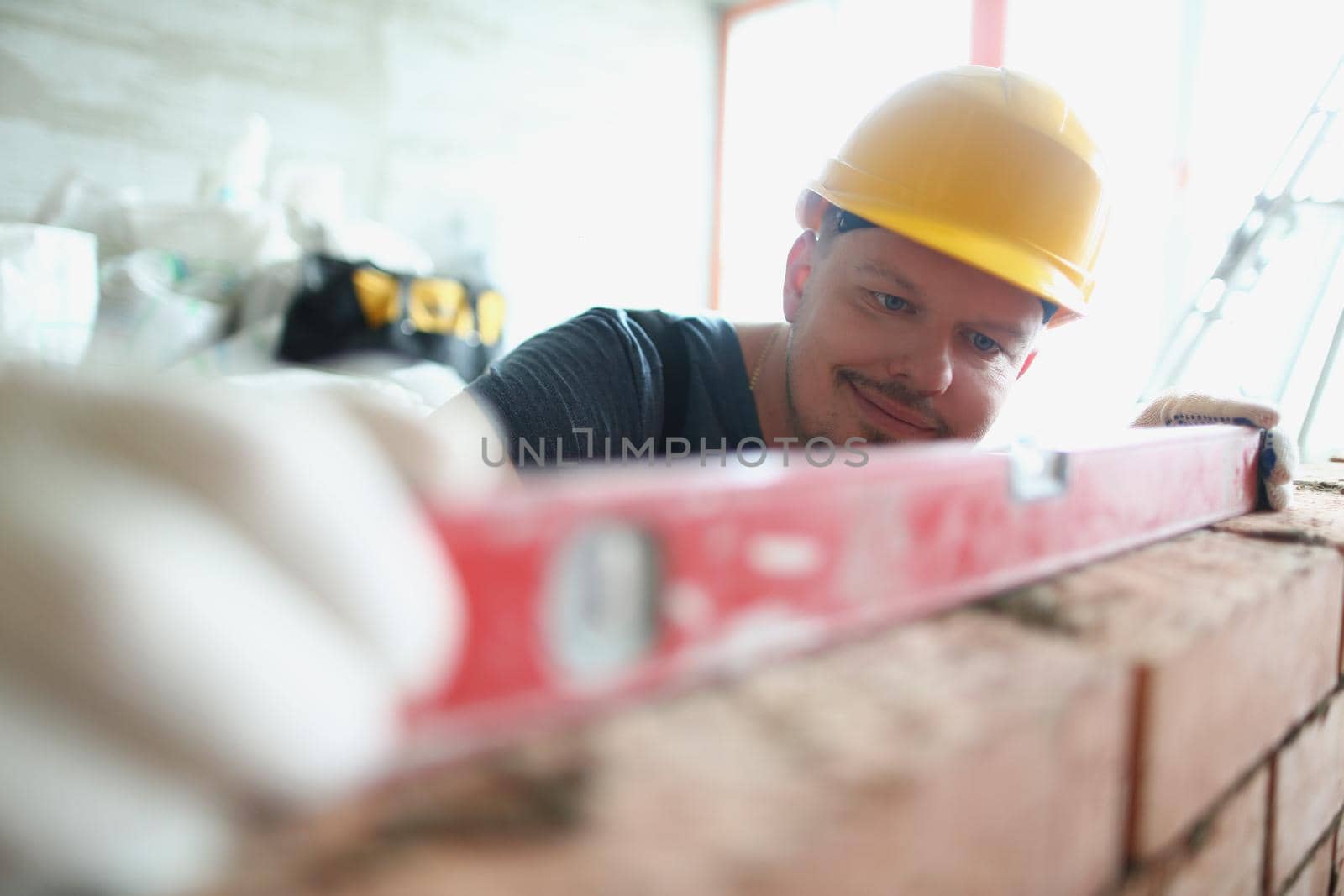 Portrait of professional male builder measure distance on wall with level tool, concrete barrier made of red bricks. Renovation, construction site concept