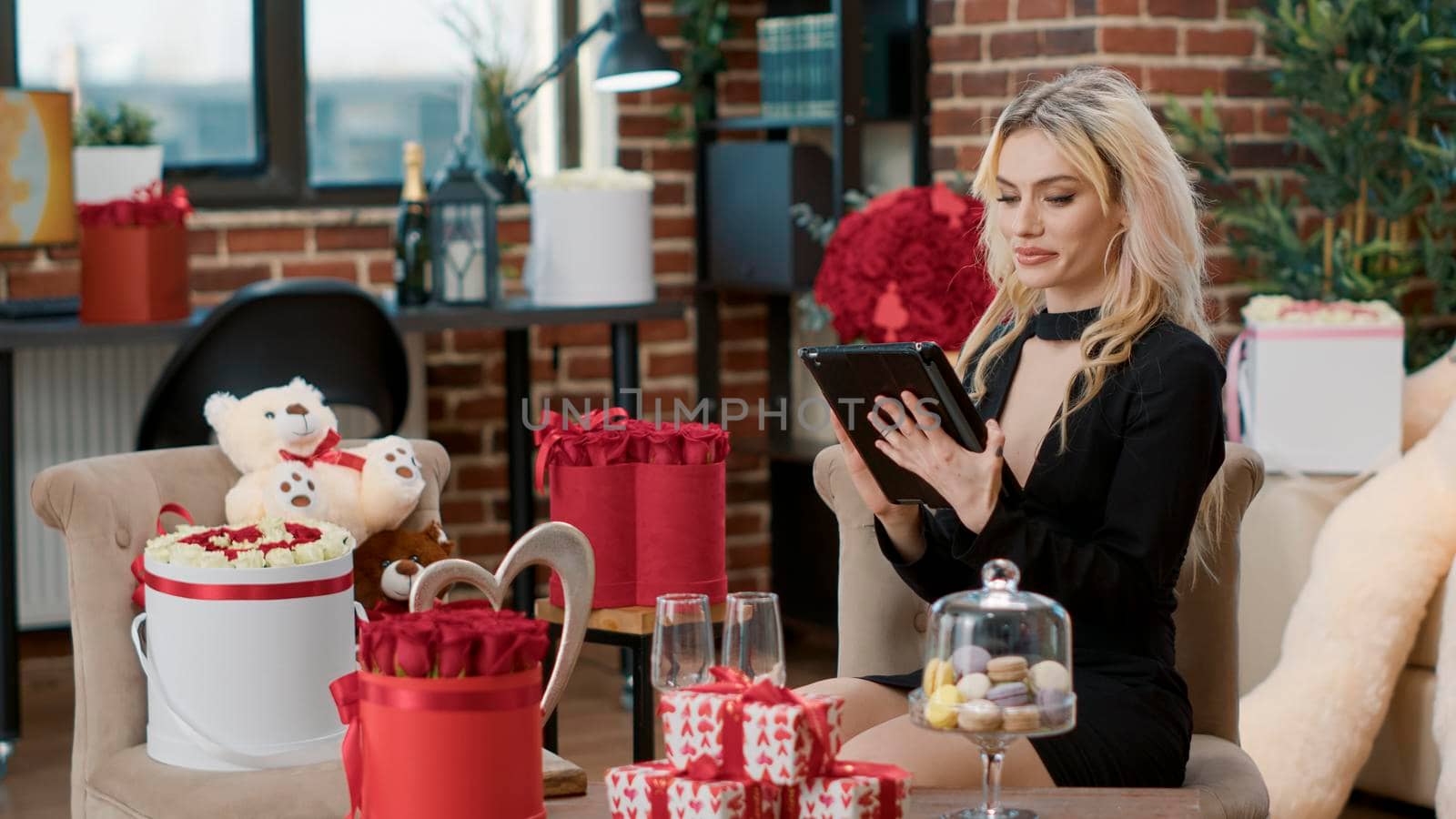 Elegant blonde woman smiling at tablet computer while talking with remote boyfriend during valentine day surprise. Attractive cute girlfriend enjoying romantic gift advertising romance anniversary