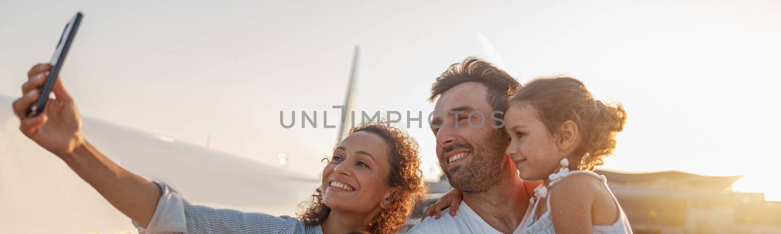 Happy tourists, beautiful family taking selfie while standing together outdoors ready for boarding the plane at sunset. Vacation, parenthood, traveling concept